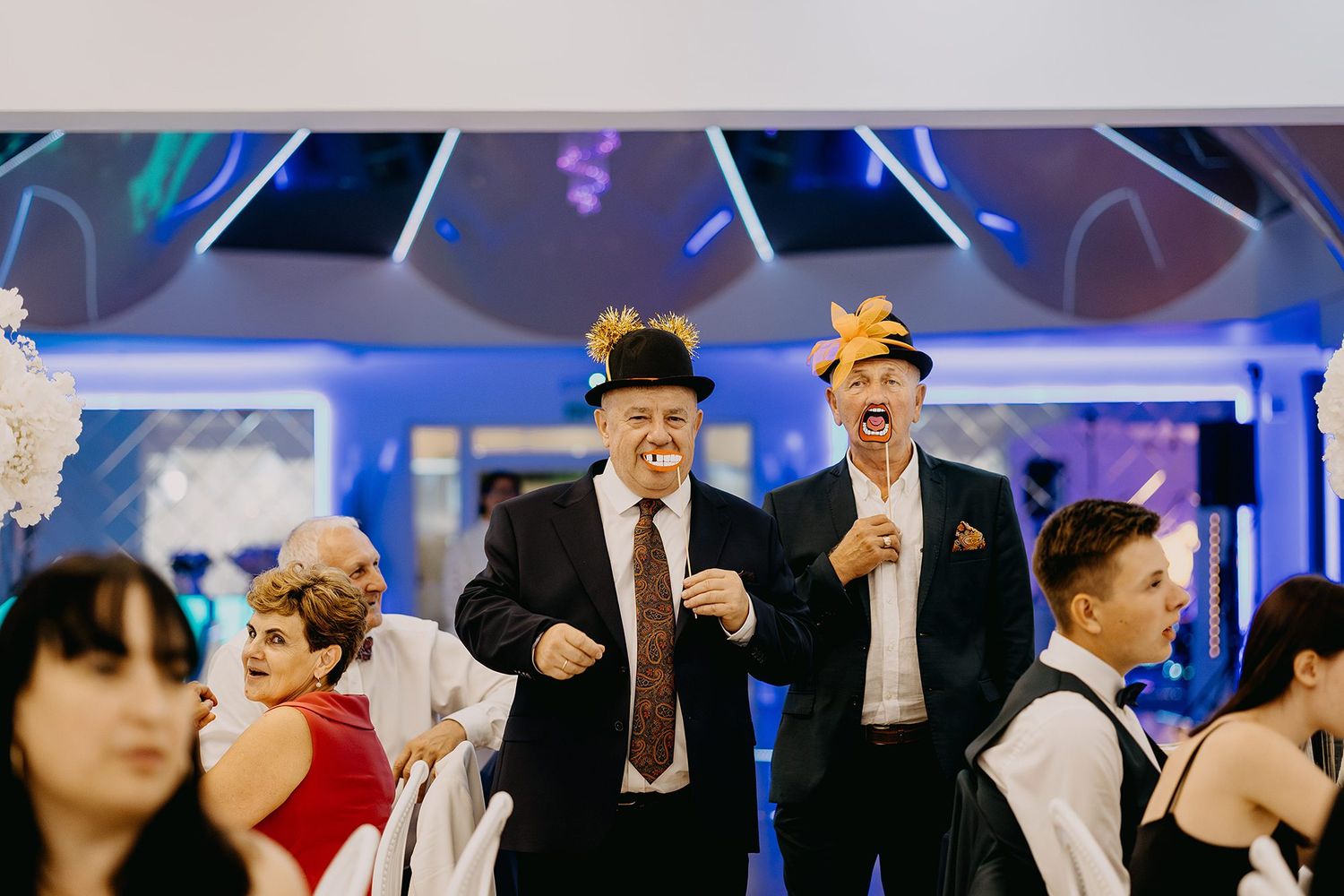 Two men in black suits and decorative hats dancing energetically at wedding reception.