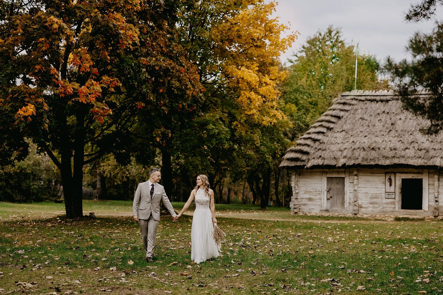 Bride and groom walking hand in hand through an autumn park near a thatched cottage.