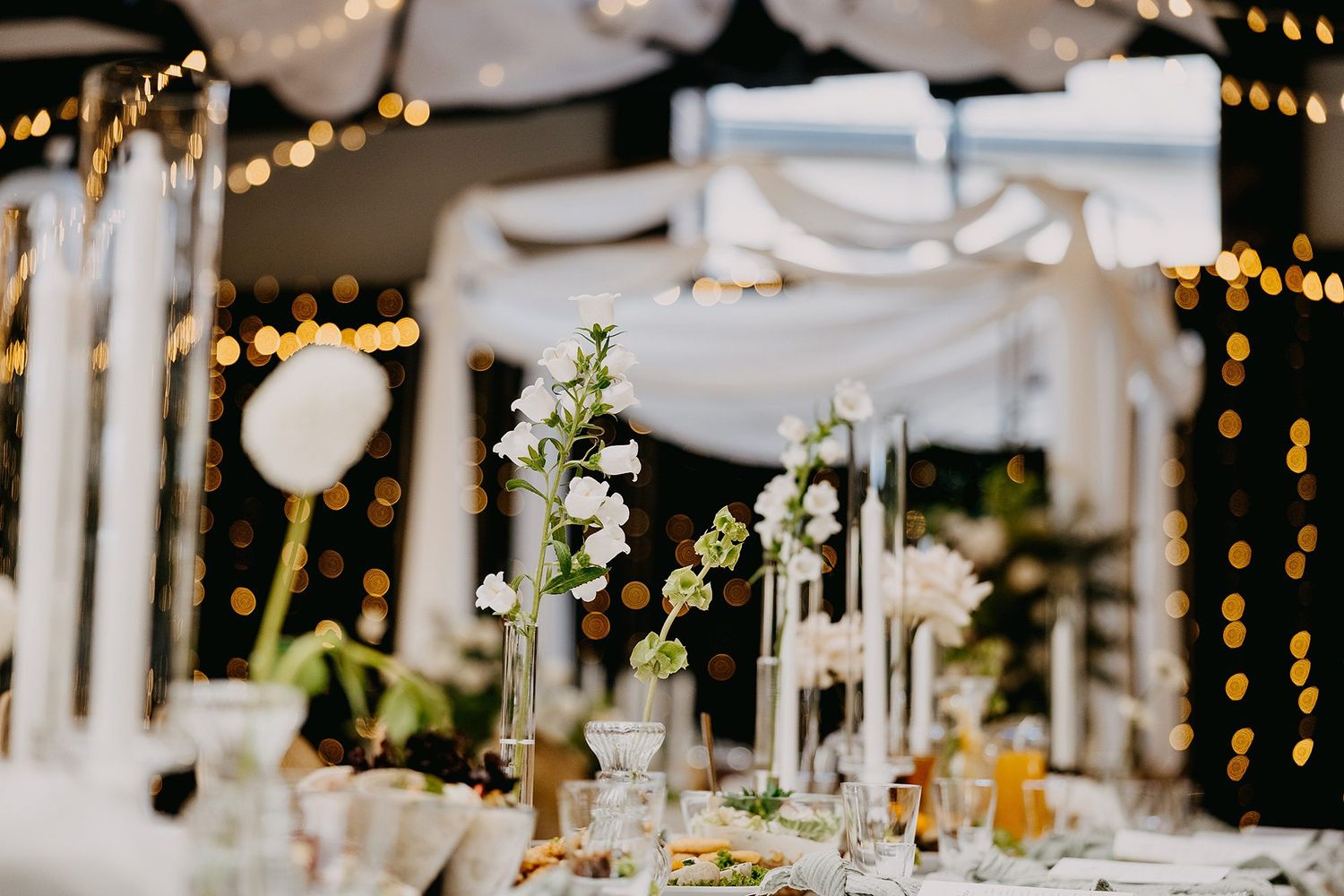 Elegant wedding reception table adorned with white floral arrangements, tall candles, and warm bokeh lights.