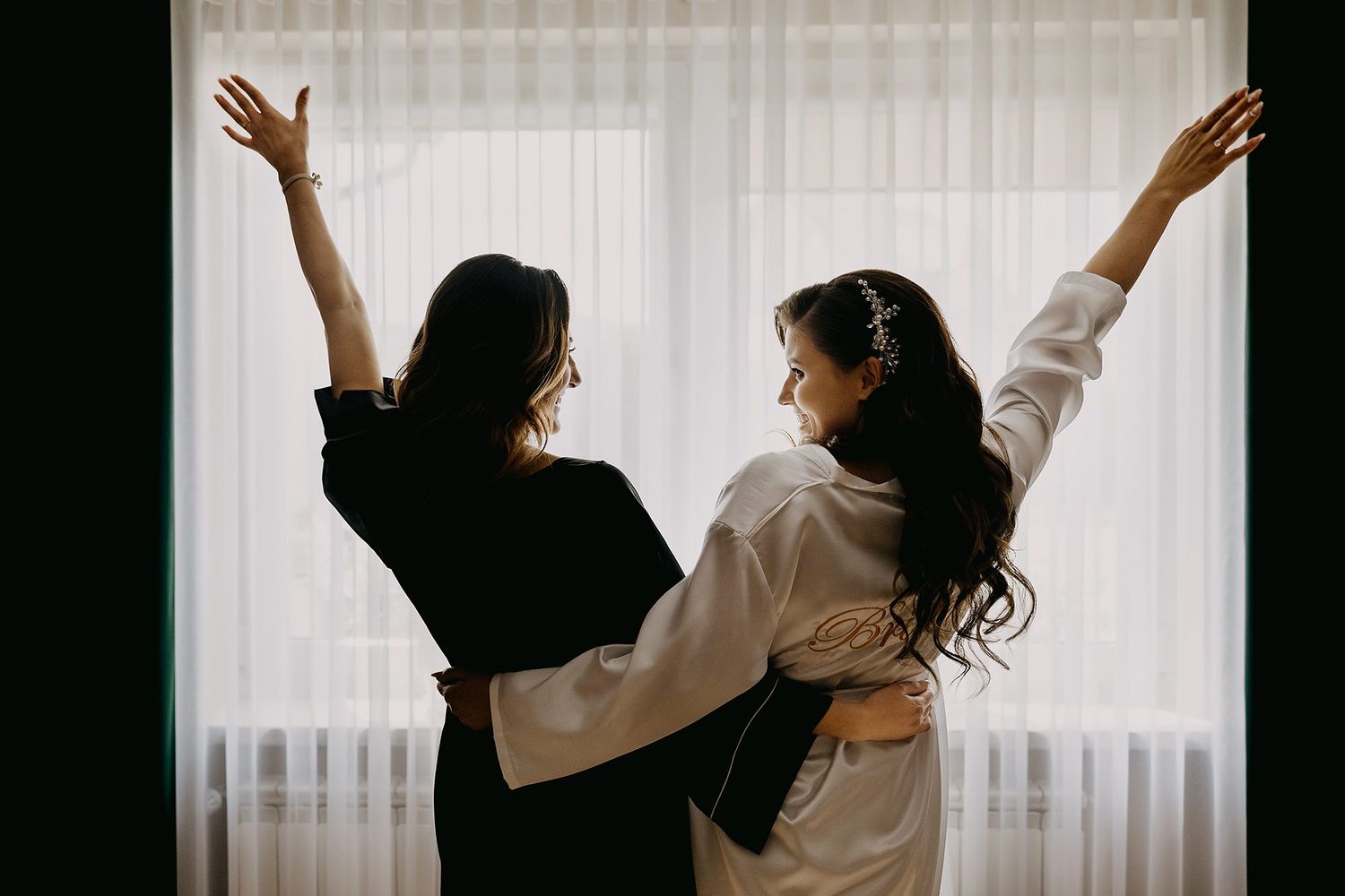 Two women in robes celebrating with arms raised by bright curtained window.