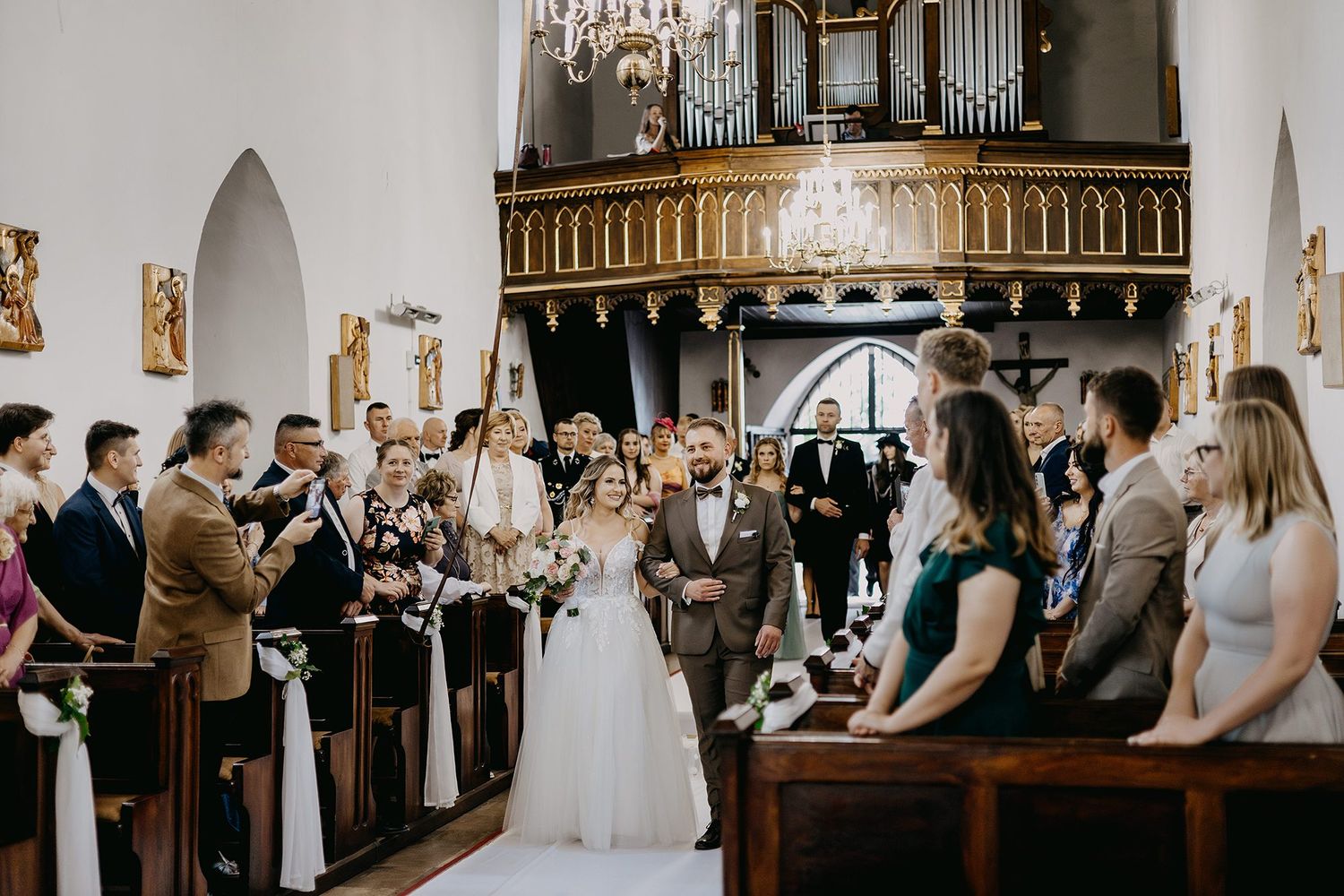 Bride walks down church aisle with escort past guests in historic church with pipe organ.