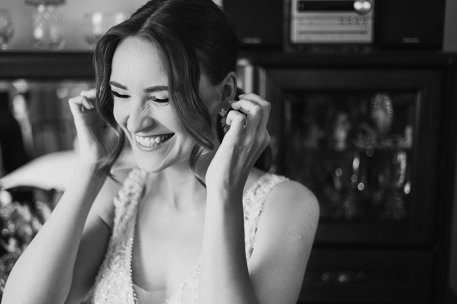 Black and white portrait of joyful bride laughing while putting on earrings before wedding.