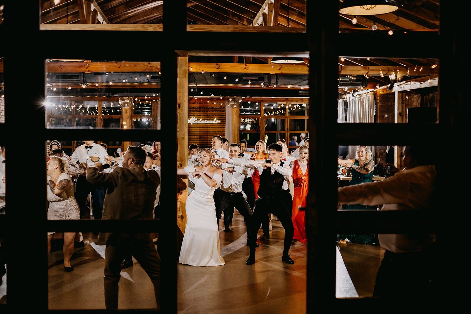 Wedding reception dance floor viewed through glass doors with festive string lights overhead.