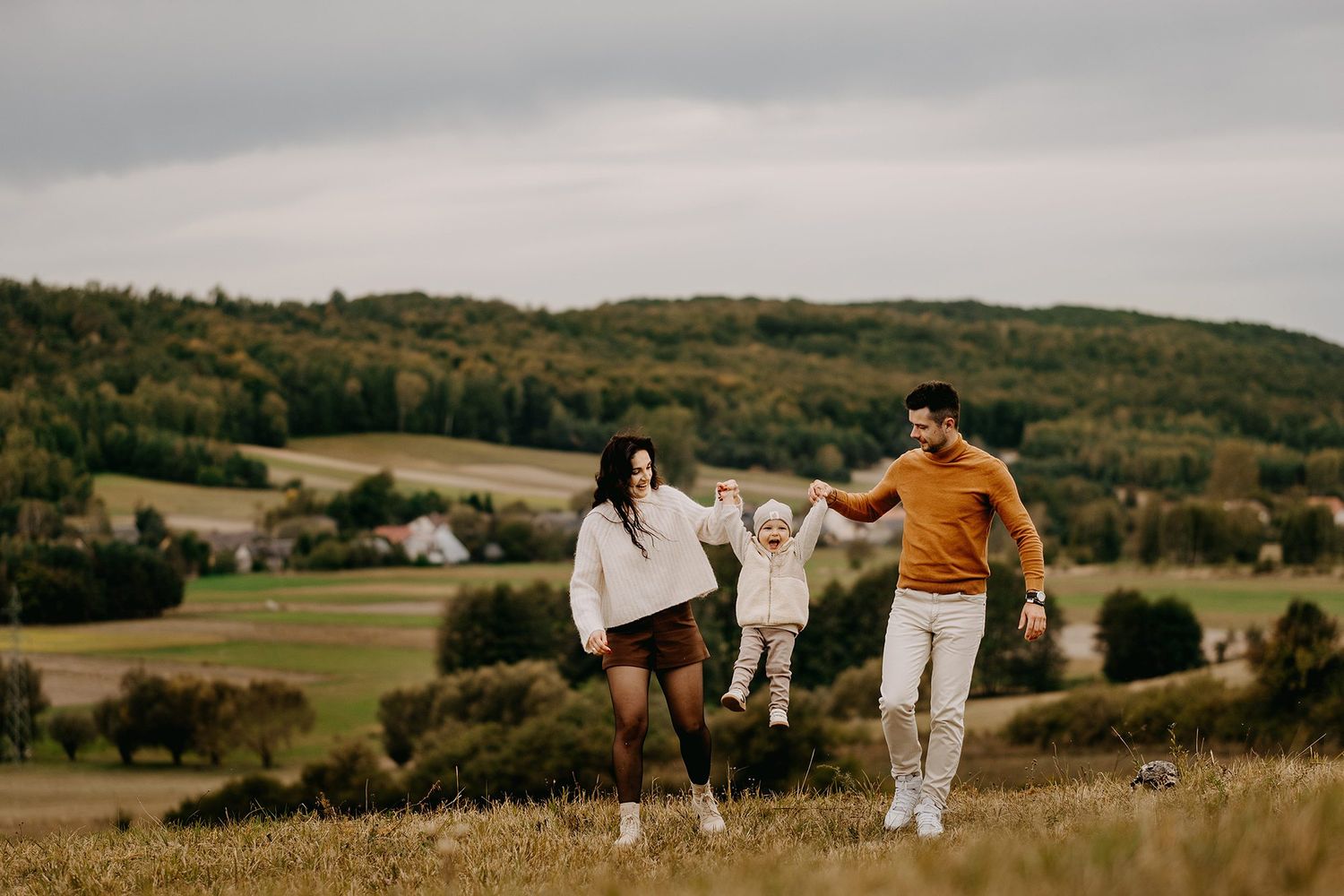 Happy couple swinging toddler between them in scenic autumn countryside field.