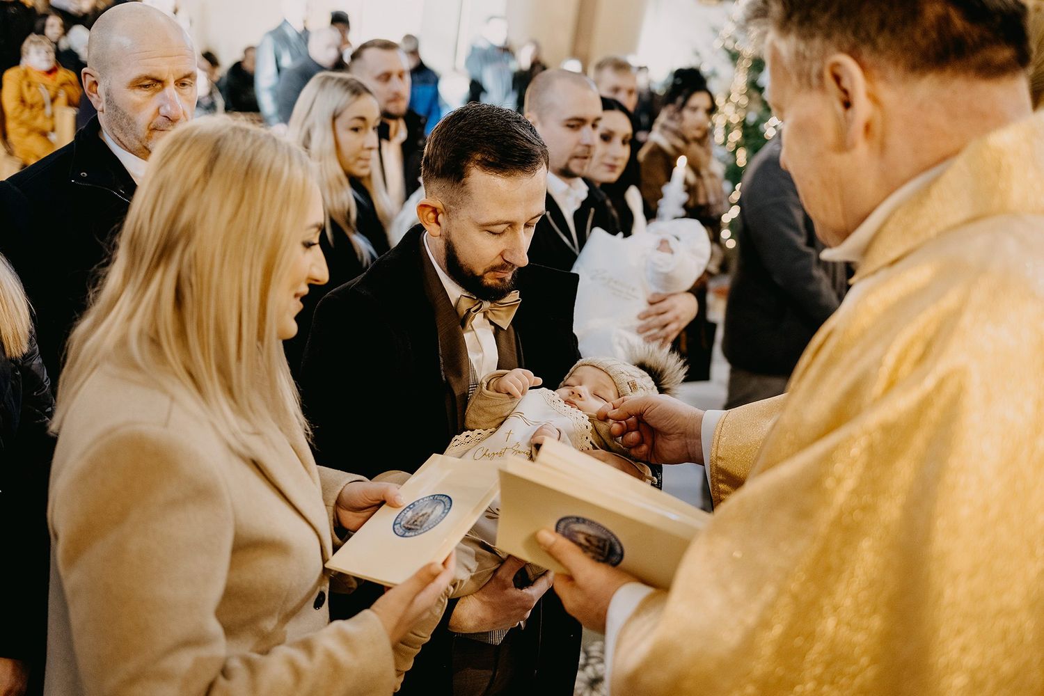 Priest blessing a baby during a christening ceremony with parents holding religious items in a church.