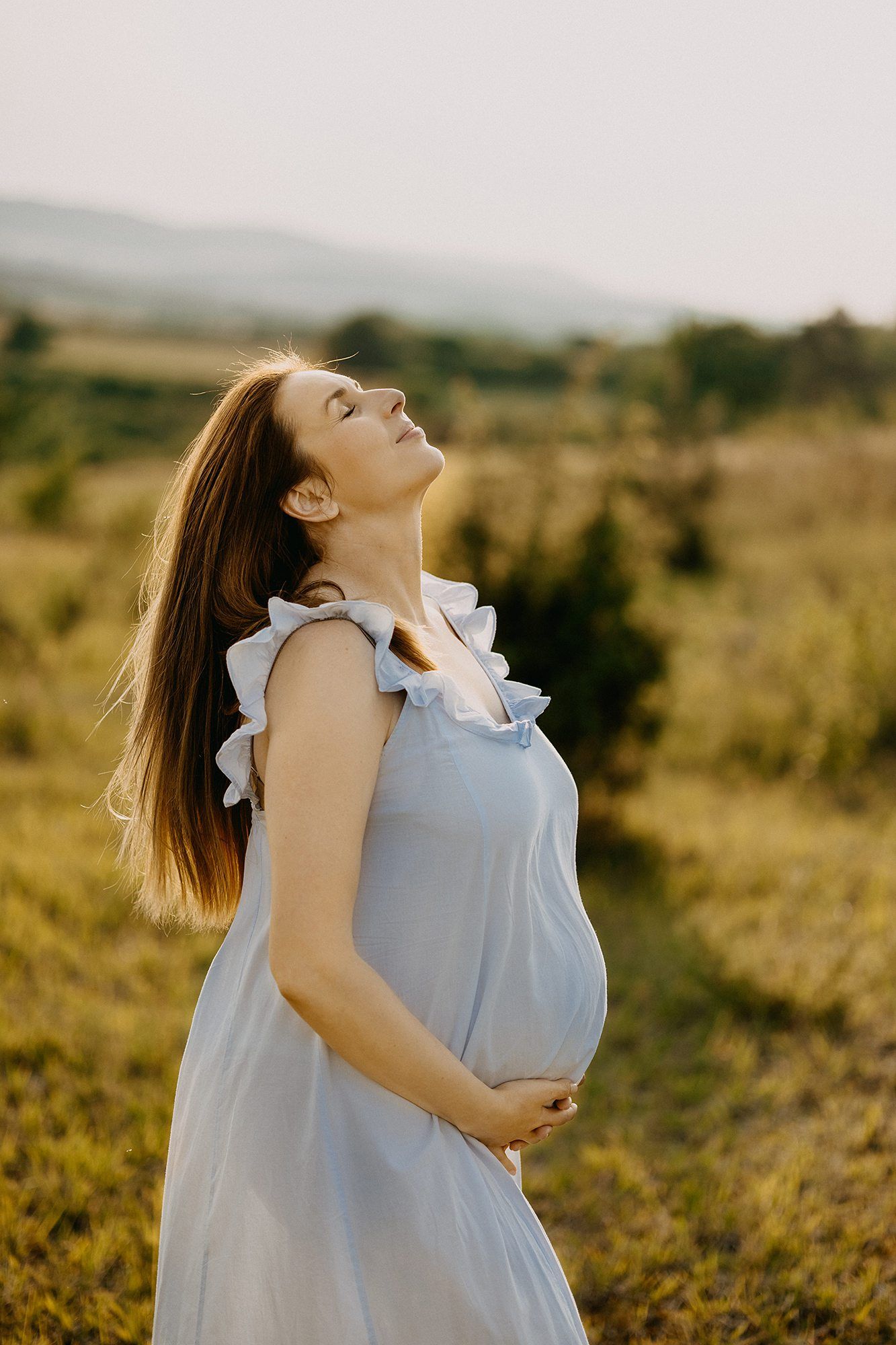 Serene pregnant woman in light blue dress holding baby bump with eyes closed in a sunny meadow.