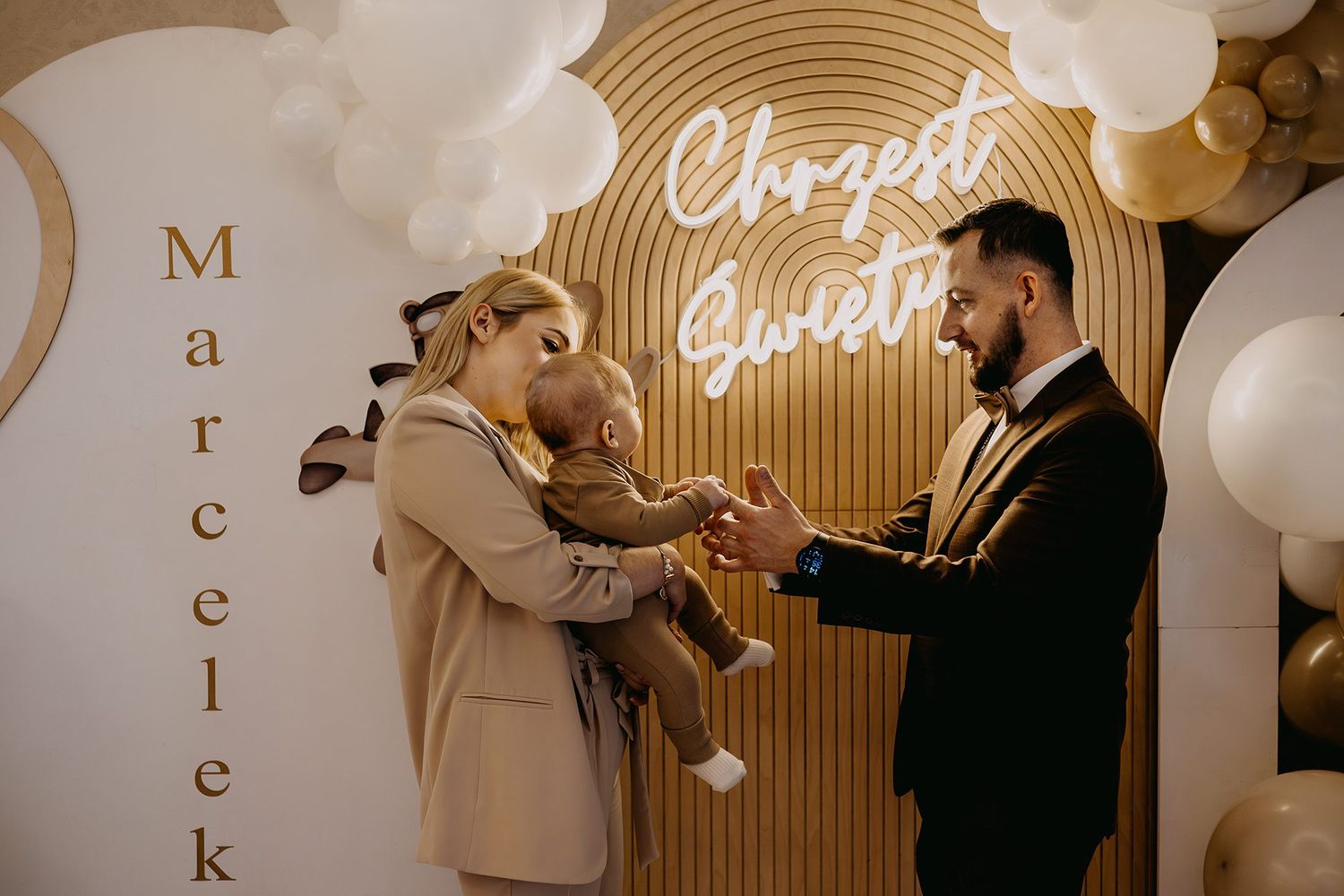 Parents present baby Marcelek at a decorated christening backdrop with balloon arch.