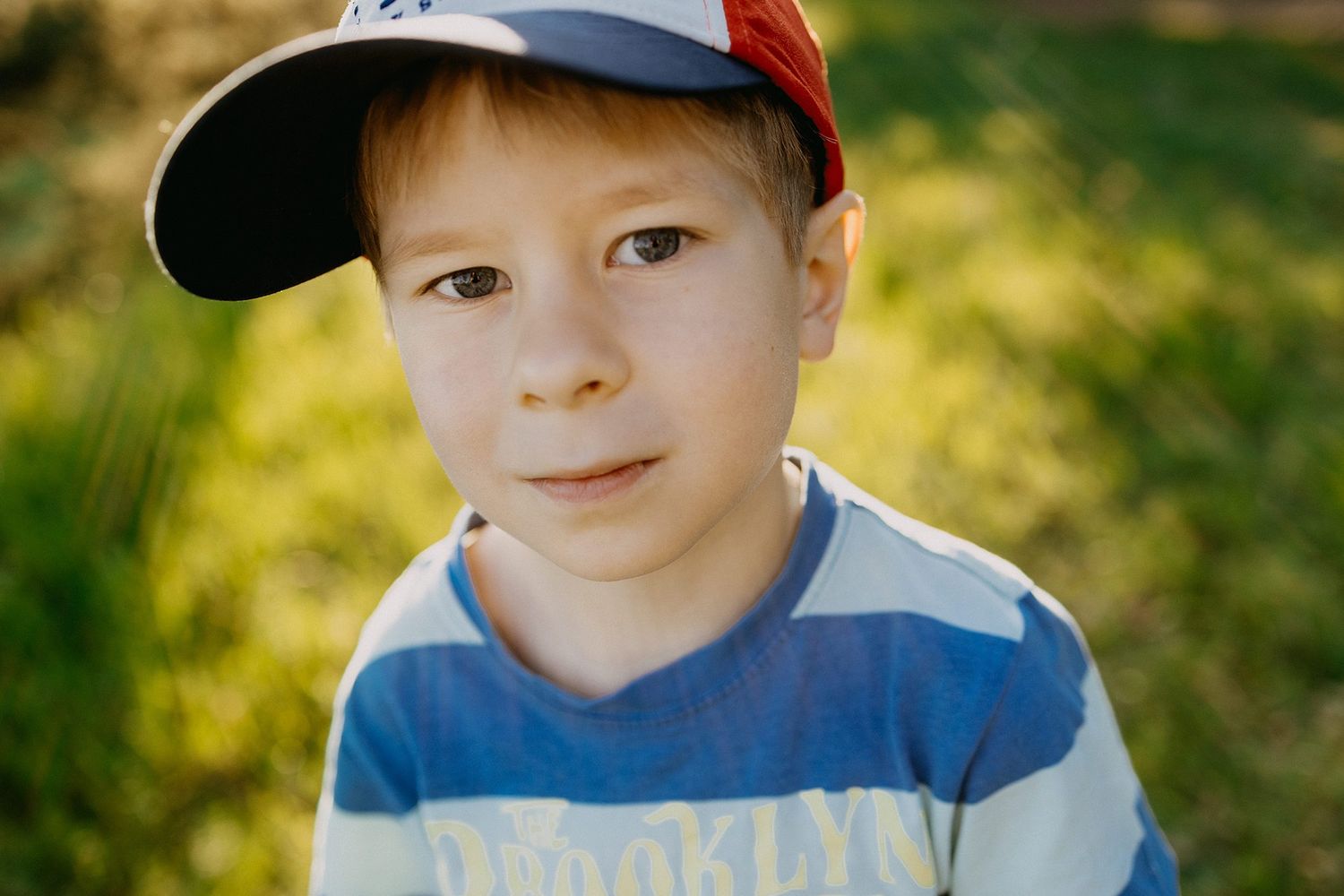 A cute young boy wearing a Brooklyn striped shirt and red baseball cap poses outdoors.