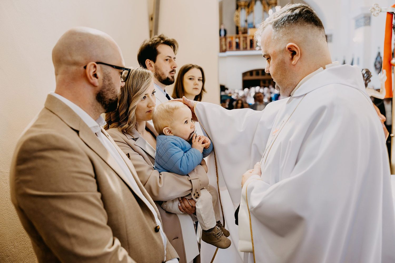 A priest performs a baptism ceremony blessing a baby held by parents in a church.