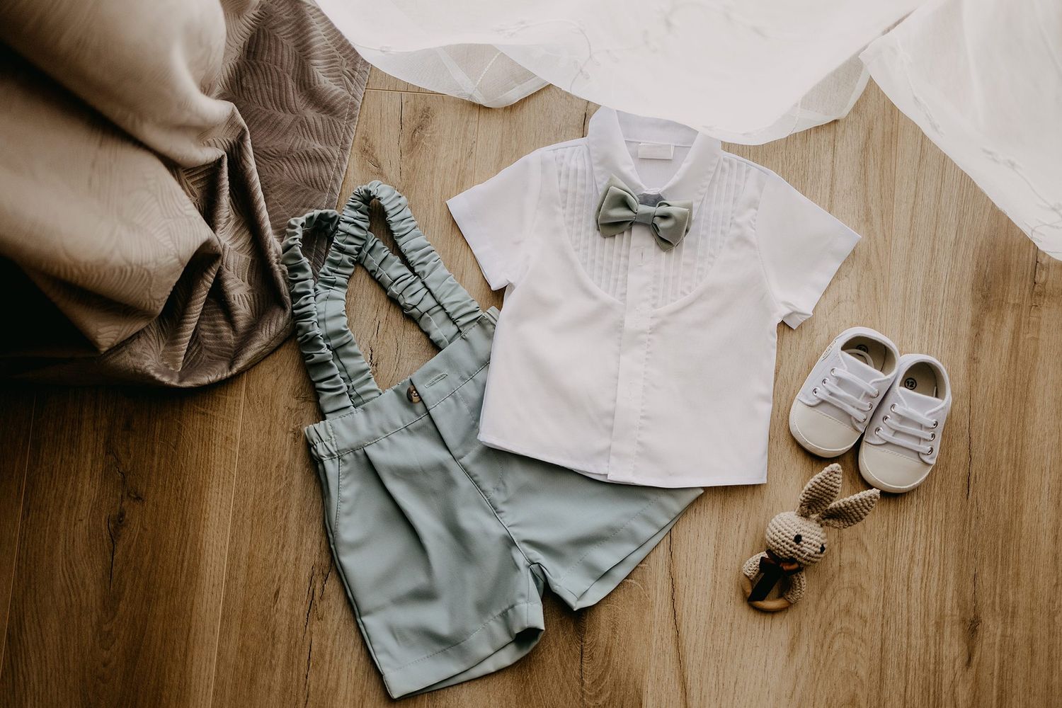 Baby boy baptism outfit with white shirt, sage suspender shorts, bow tie, and white shoes on wood floor.