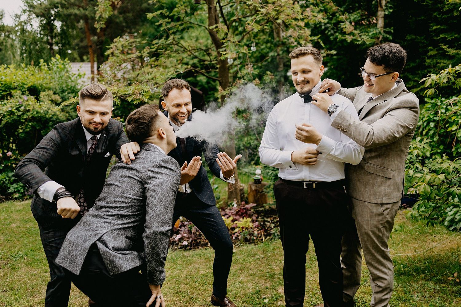 Groomsmen in suits celebrating outdoors, laughing and having fun together in a garden setting on a wedding day.