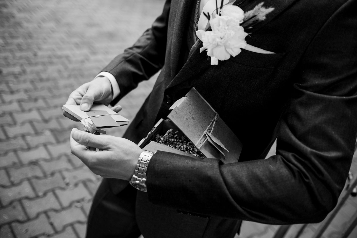 Groom in black suit holding a gift box with white boutonniere, black and white wedding photo.