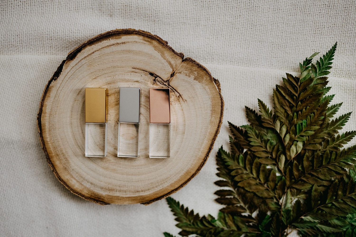 Four metallic USB drives in gold, gray, and pink on wooden slice with green fern leaves.