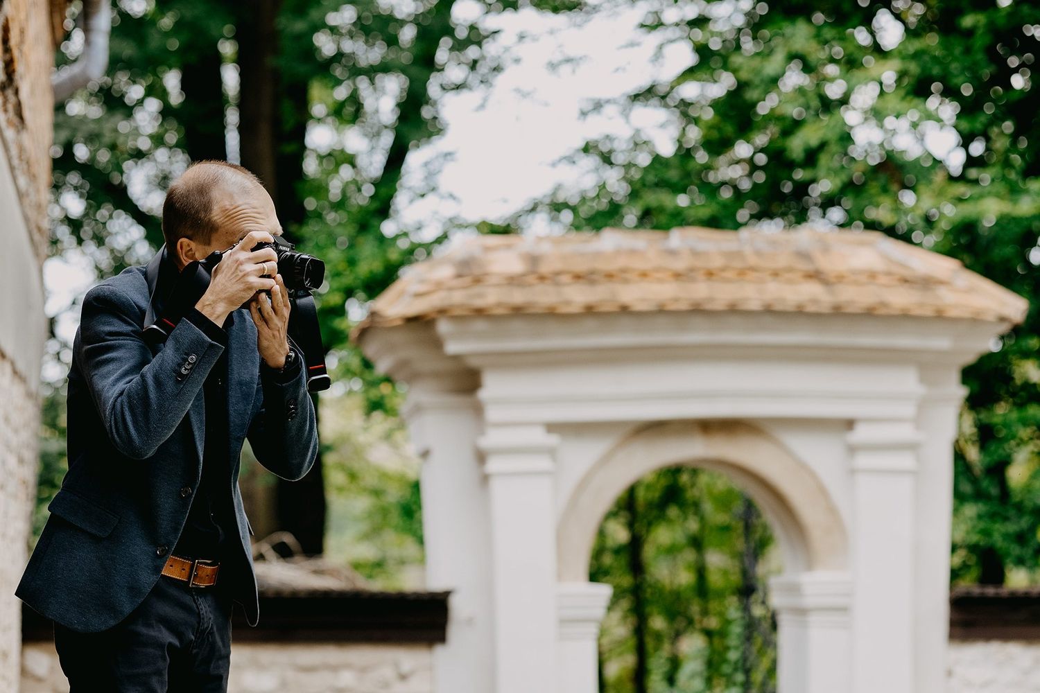 Elegant male photographer in suit capturing images near ornate white garden gate in forest.