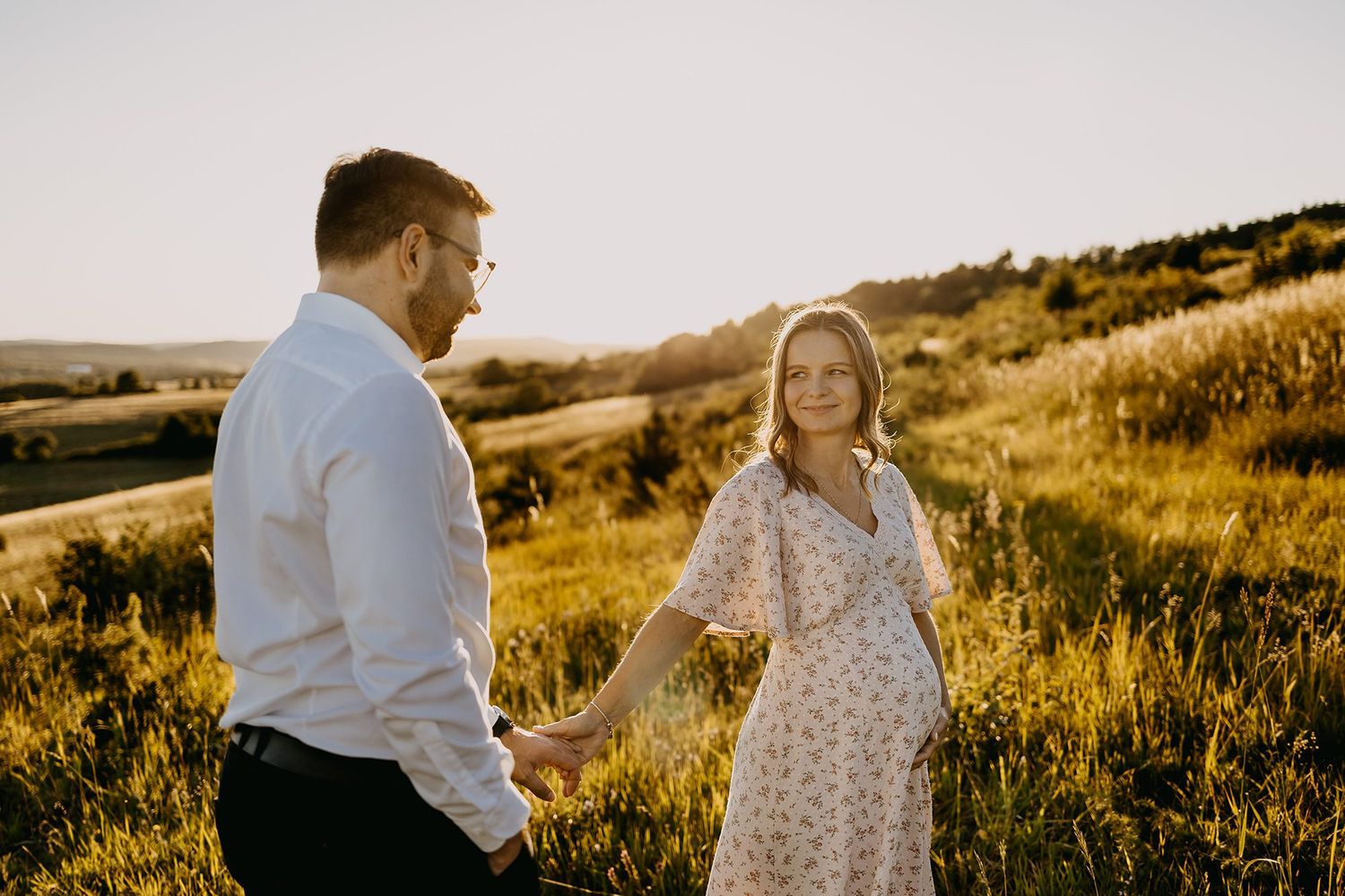 Pregnant couple holding hands in a sunlit meadow, woman in floral dress smiling at partner.