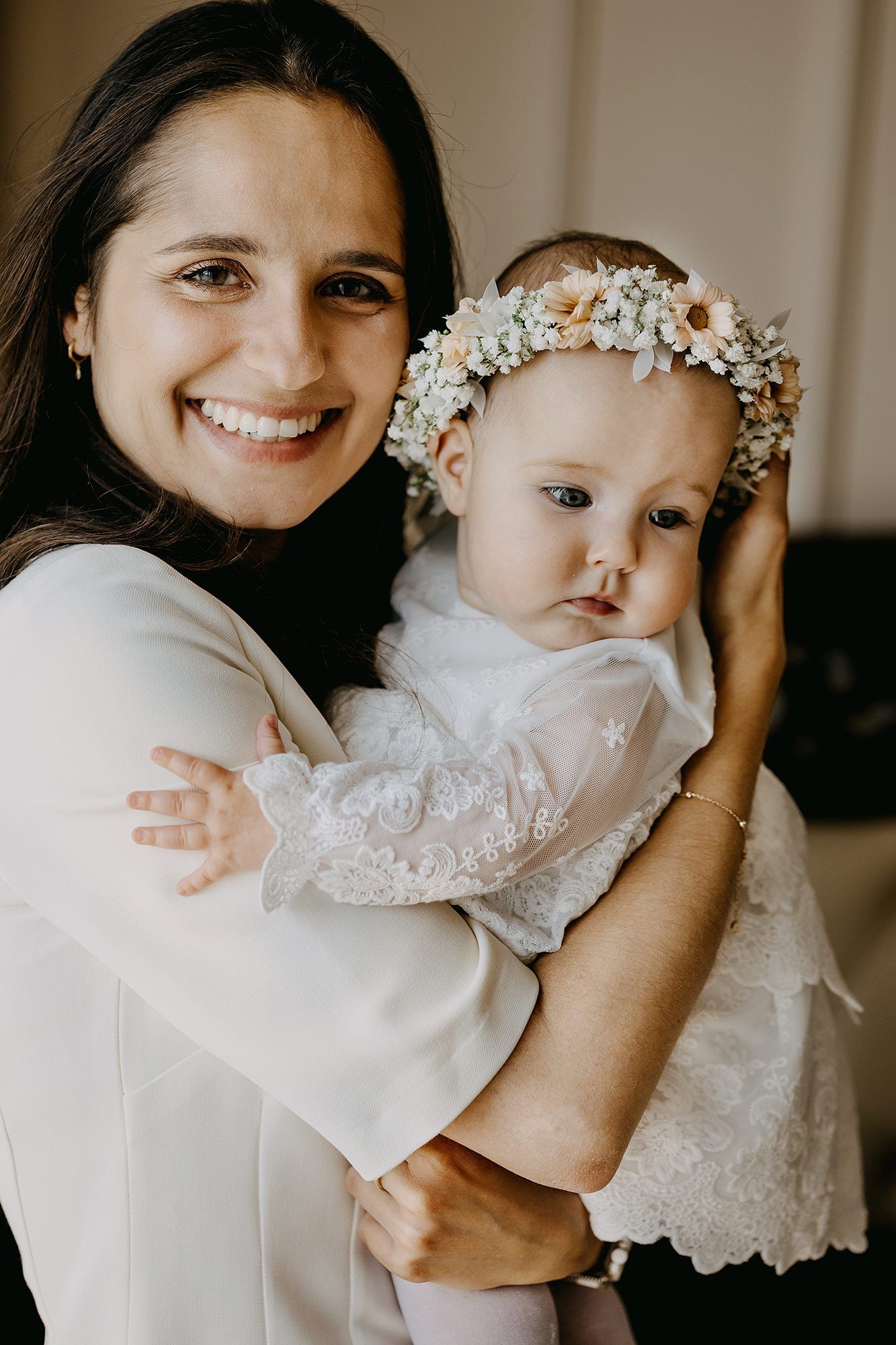 Smiling mother holding baby girl wearing white lace christening dress and floral flower crown headband.