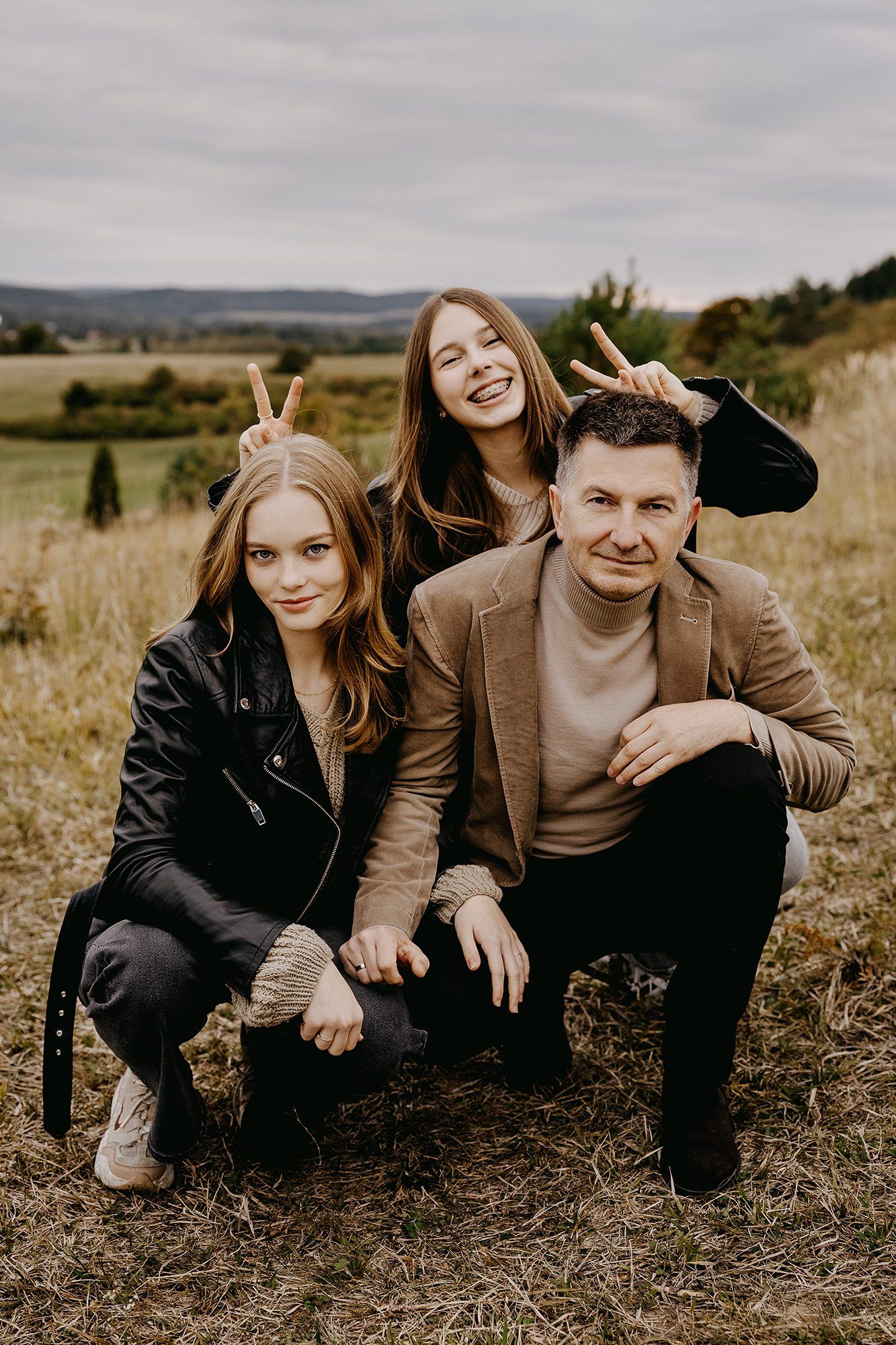 Father and two teenage daughters posing playfully with bunny ears in an autumn field.