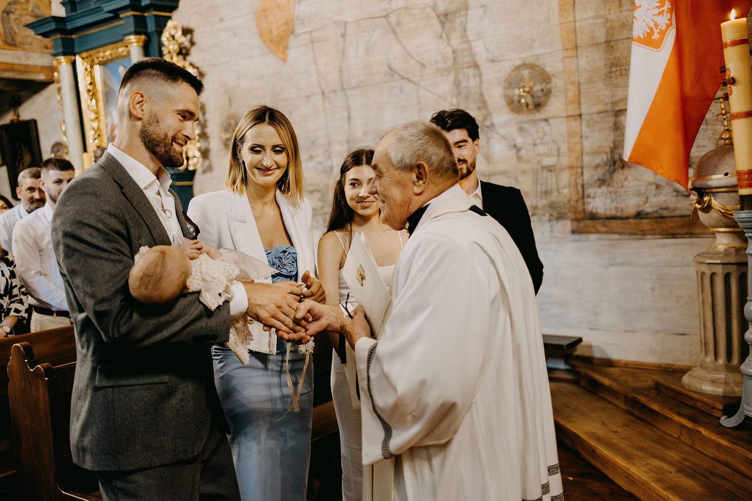 Smiling family presents newborn baby to a priest during a Catholic baptism ceremony inside a historic church.