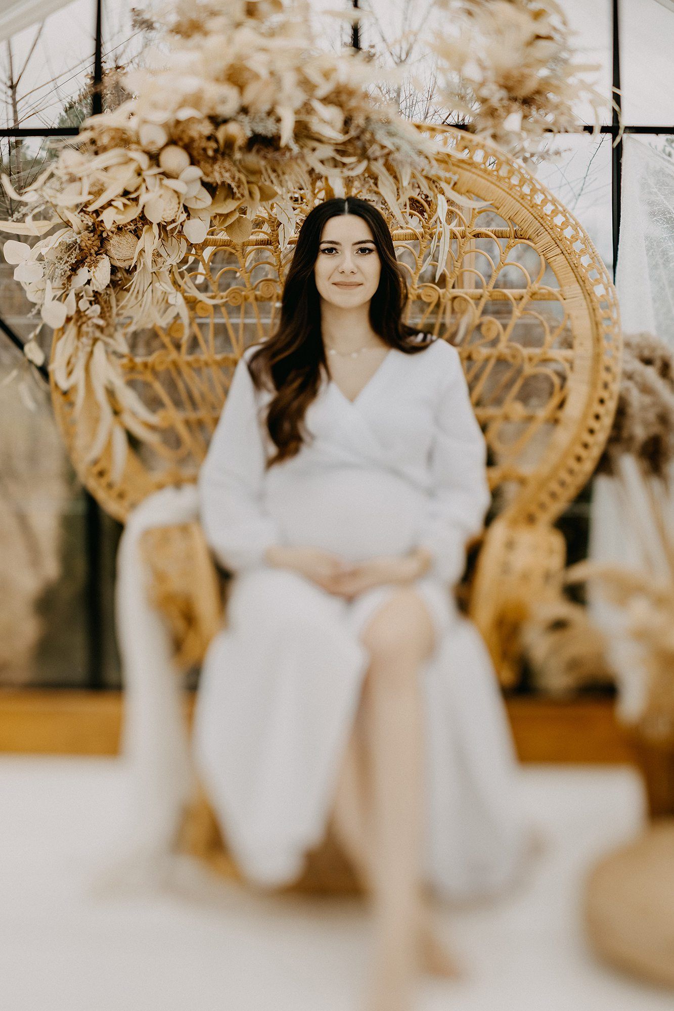 Elegant pregnant woman in white dress sitting in rattan peacock chair surrounded by dried floral decor.