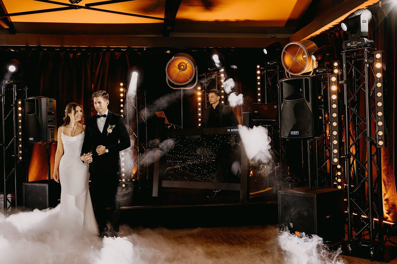 Bride and groom walk through dramatic fog and stage lights at their elegant wedding reception.