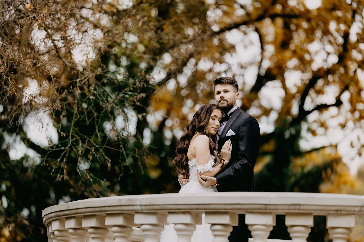 Bride and groom share close embrace on stone balustrade surrounded by golden autumn trees.