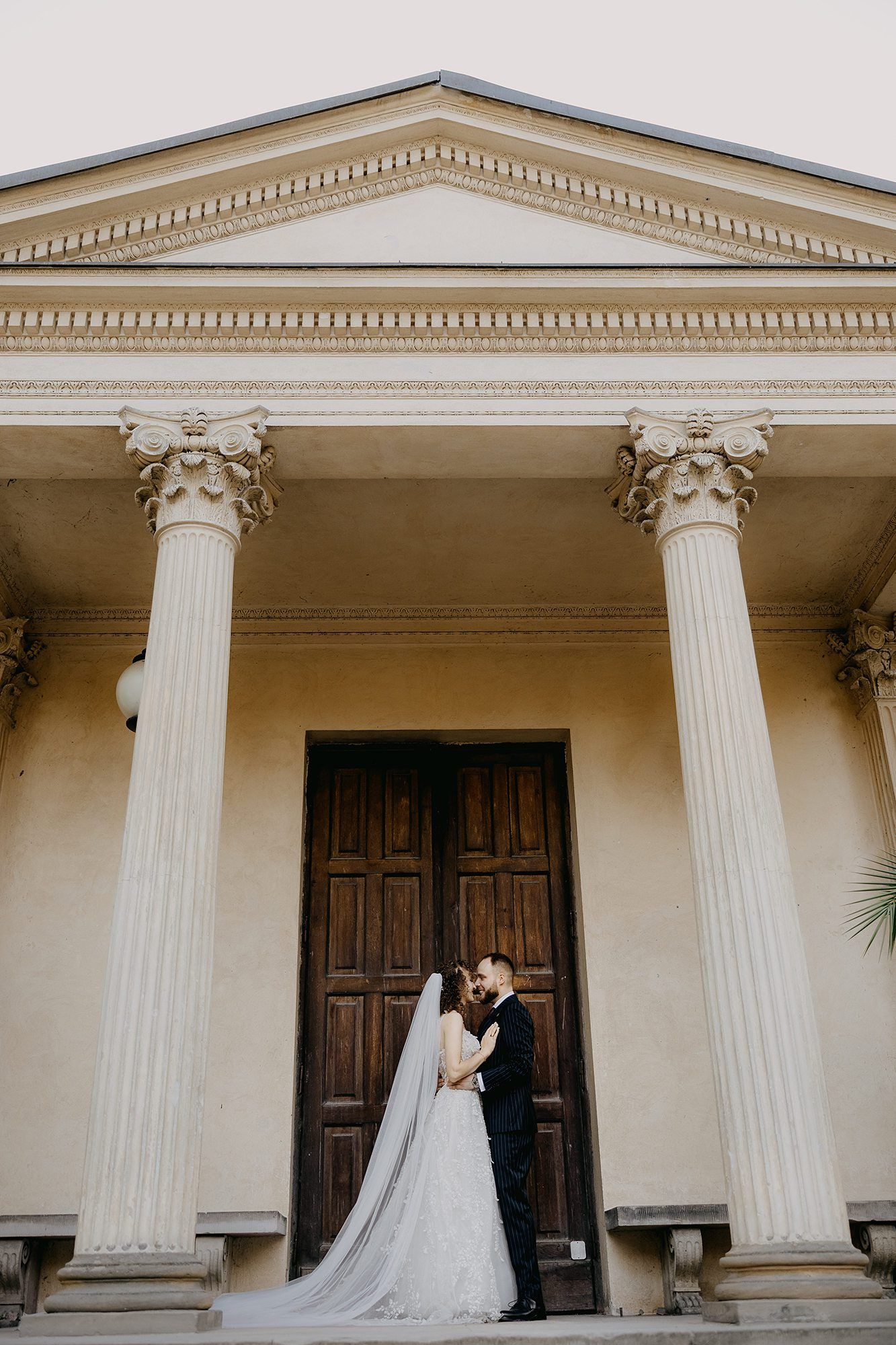 Bride and groom kiss before grand neoclassical building with tall Corinthian columns and wooden doors.