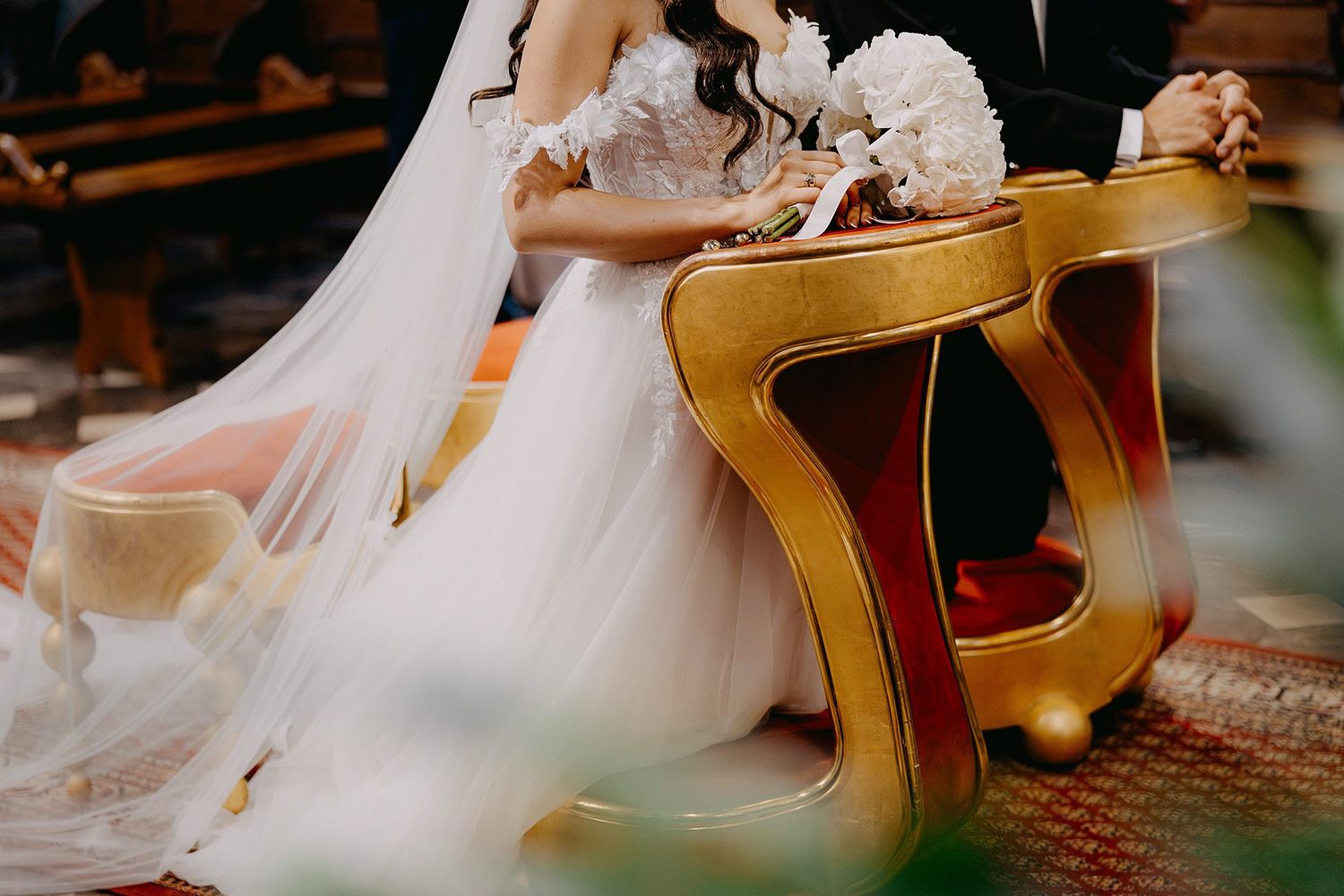 Bride in white lace wedding gown seated at ornate gold altar table during church ceremony.