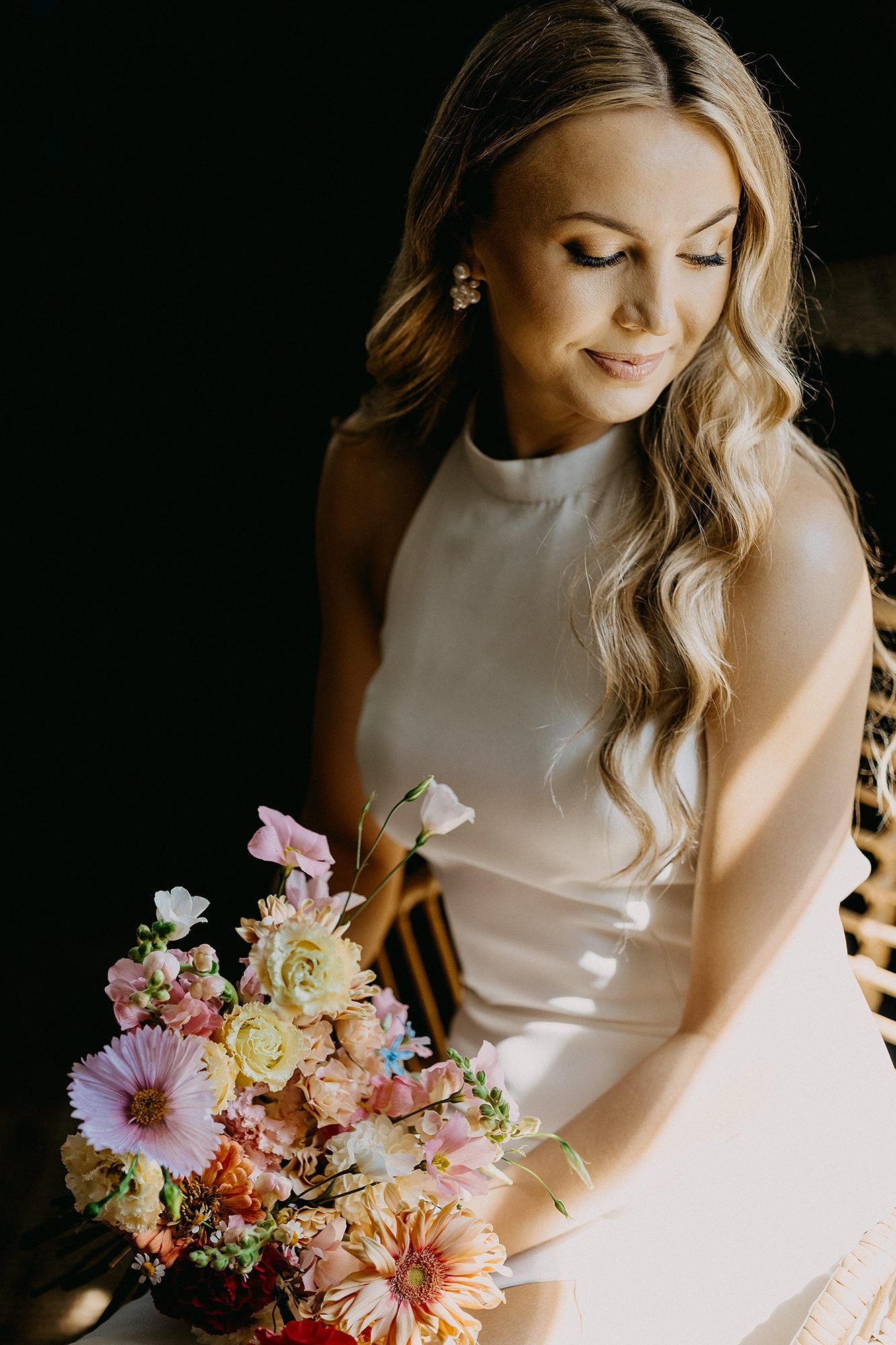 A blonde bride in a white halter gown looks down at her colorful bridal bouquet of mixed flowers.