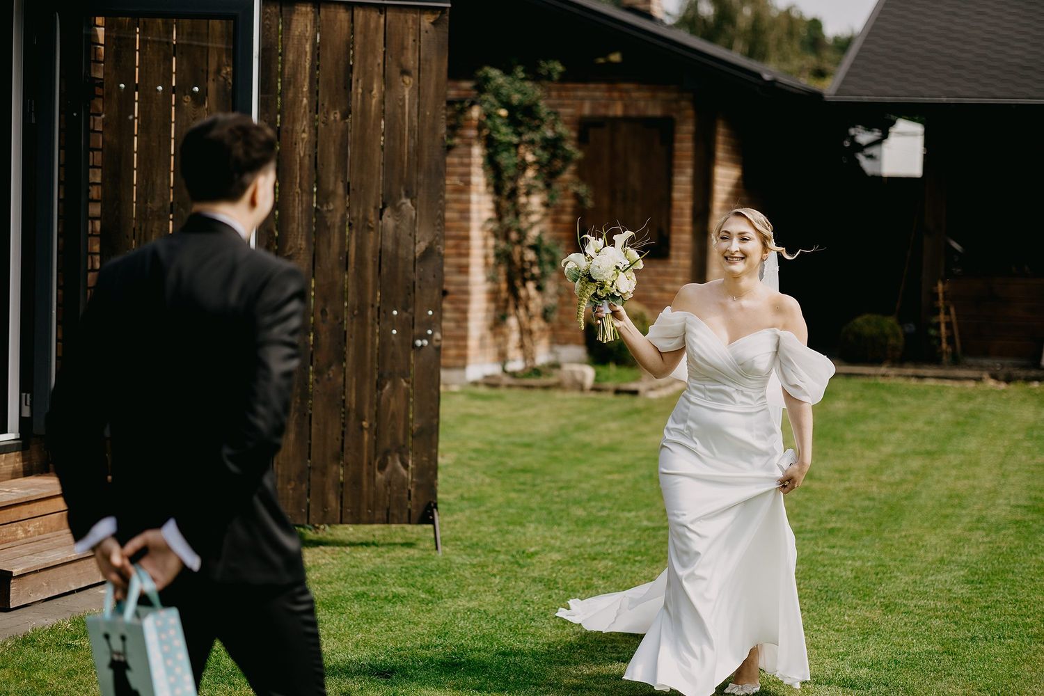 Smiling bride in white gown carries bouquet walking toward groom on green lawn.
