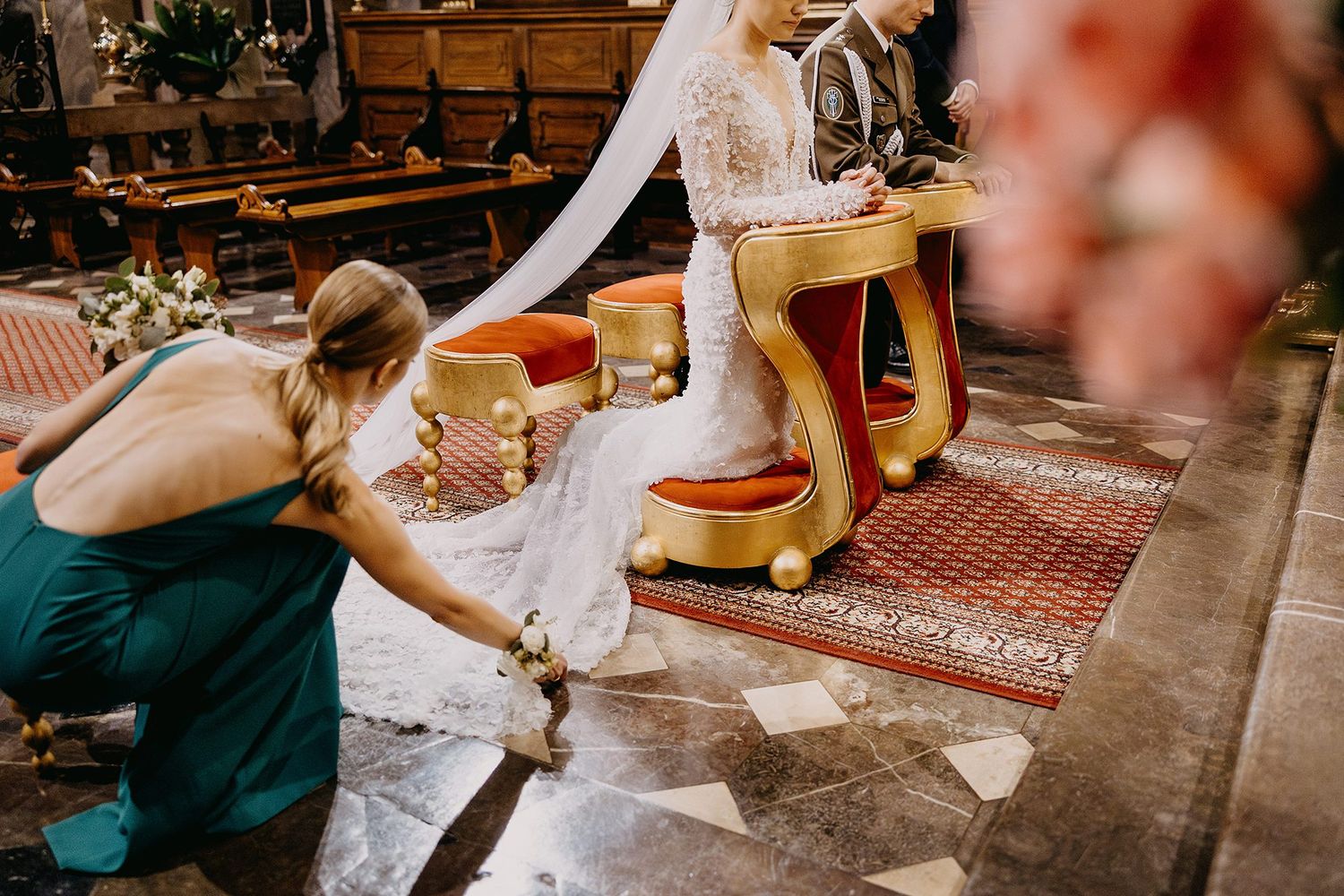 Bridesmaid in green dress adjusts the brides lace gown train during the church wedding ceremony.