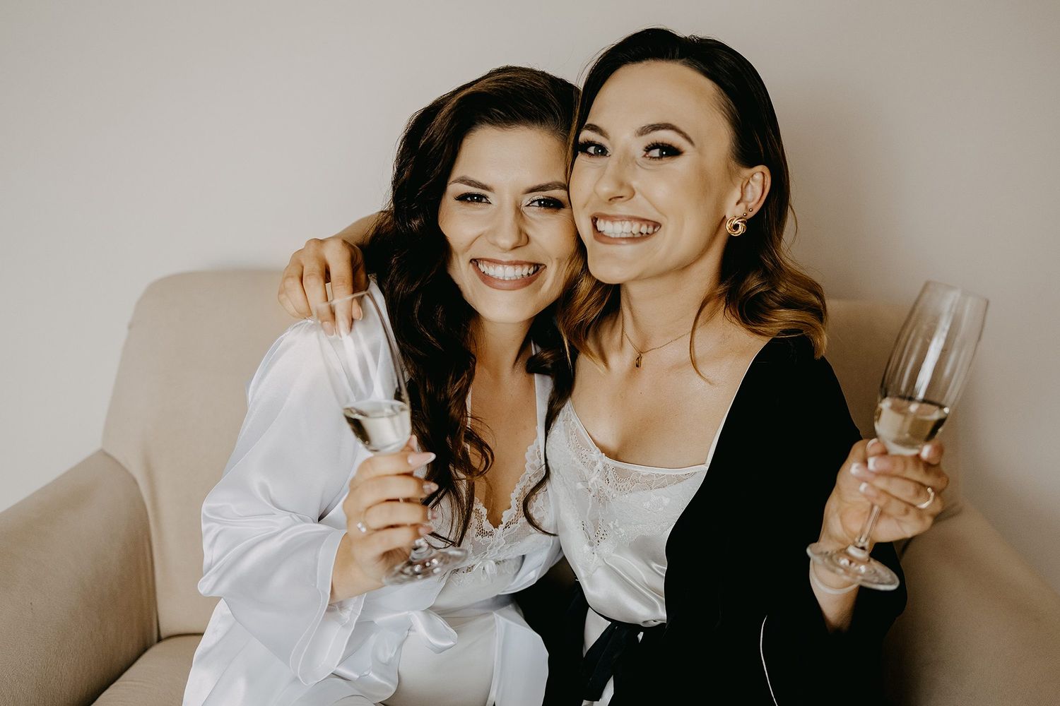 Two smiling women in robes toasting with champagne glasses while sitting together on a sofa.