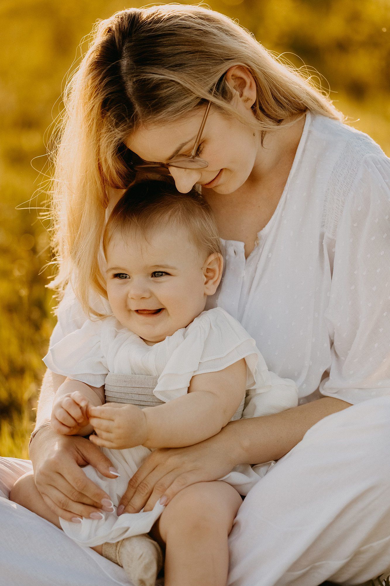 Blonde mother holding smiling baby girl in golden field.