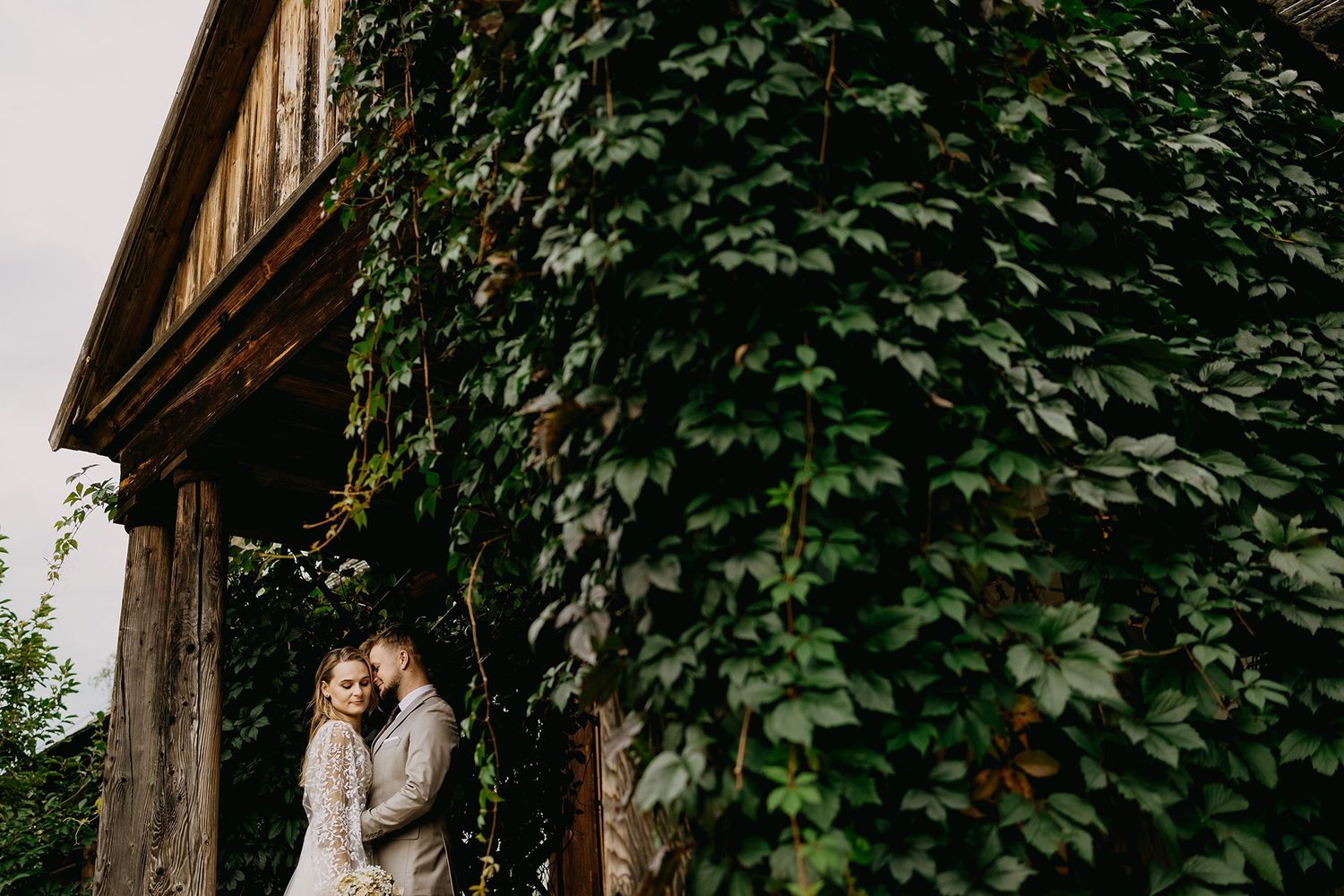 Couple embracing by ivy-covered rustic building on their wedding day.