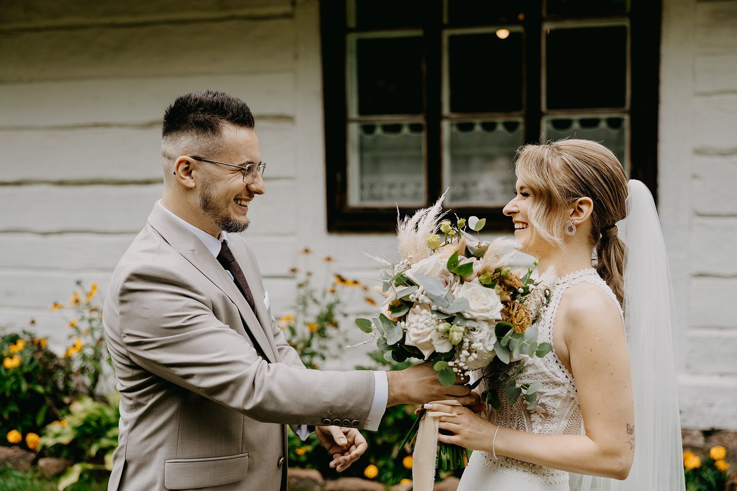 Groom in gray suit presents bride with white bouquet during outdoor wedding first look moment.