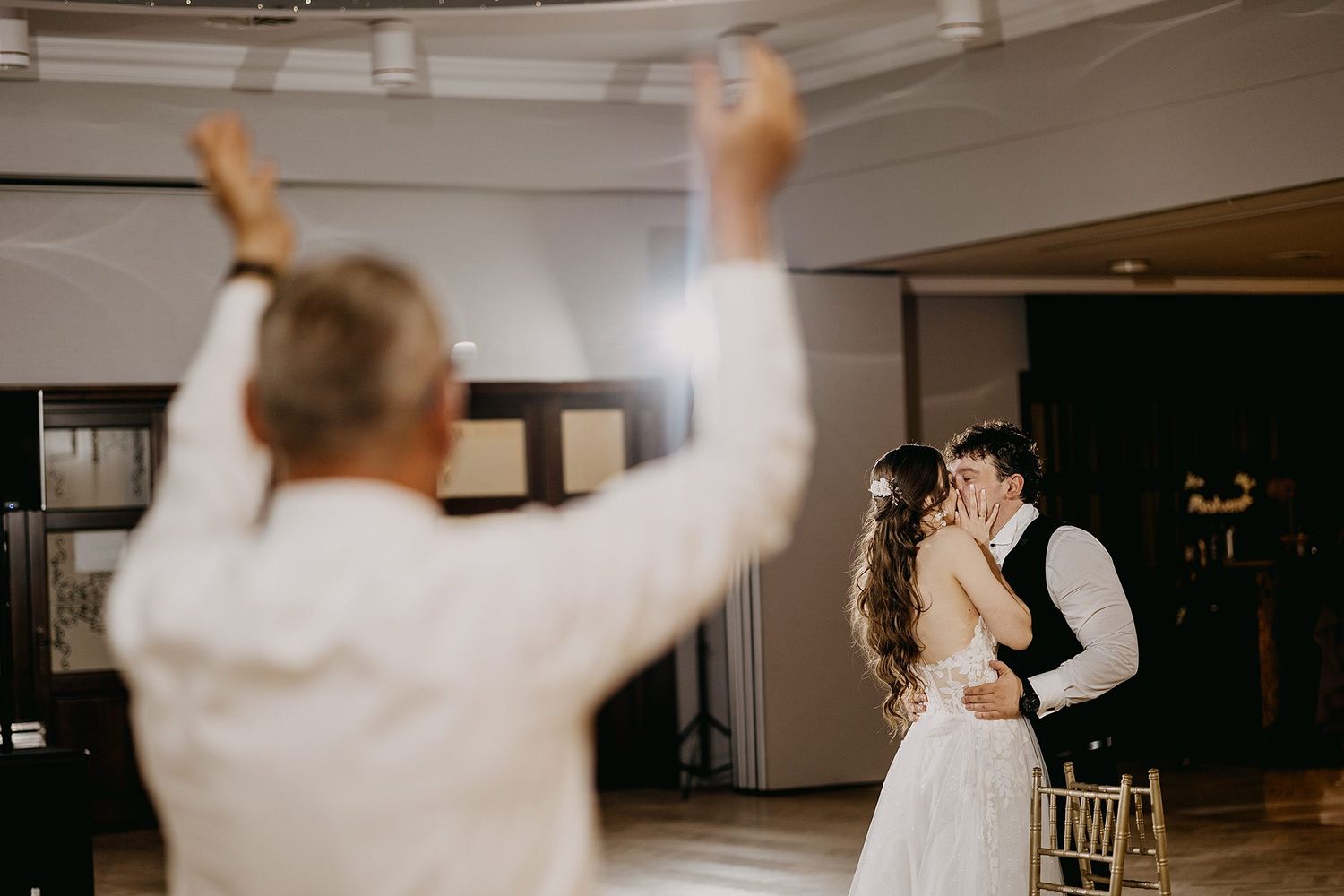 Couple shares first dance kiss as guest celebrates with raised arms at wedding reception.
