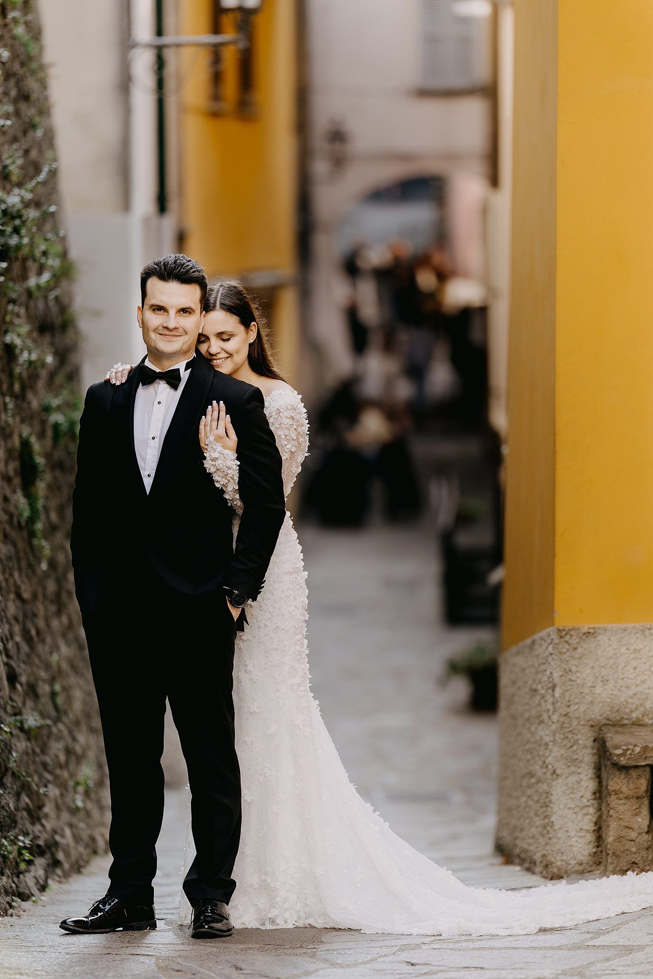Smiling groom in black tuxedo poses with bride hugging him from behind in an Italian alleyway.