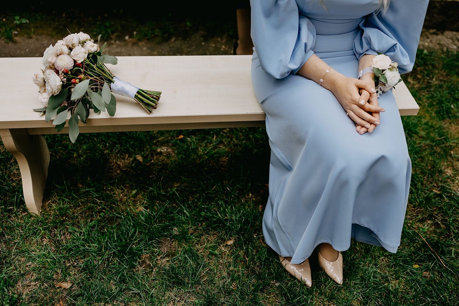 Bridesmaid in blue dress sits on wooden bench beside a white and pink wedding bouquet outdoors.