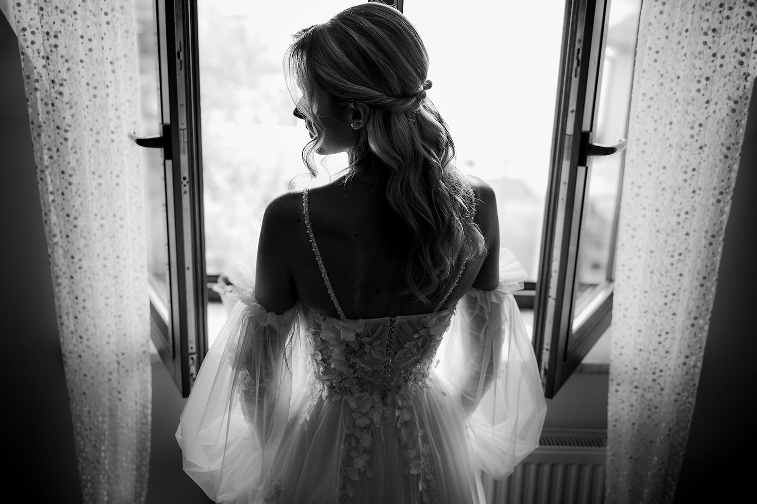 Bride in lace wedding gown with bell sleeves standing at open window in black and white photo.