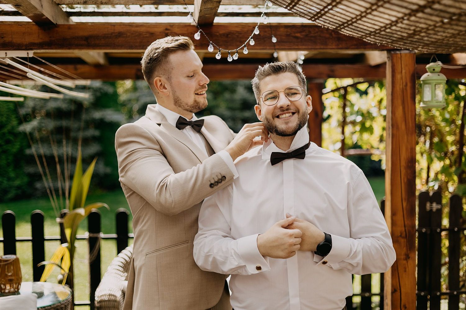 Best man adjusting groom's bow tie outdoors under a pergola before the wedding ceremony.