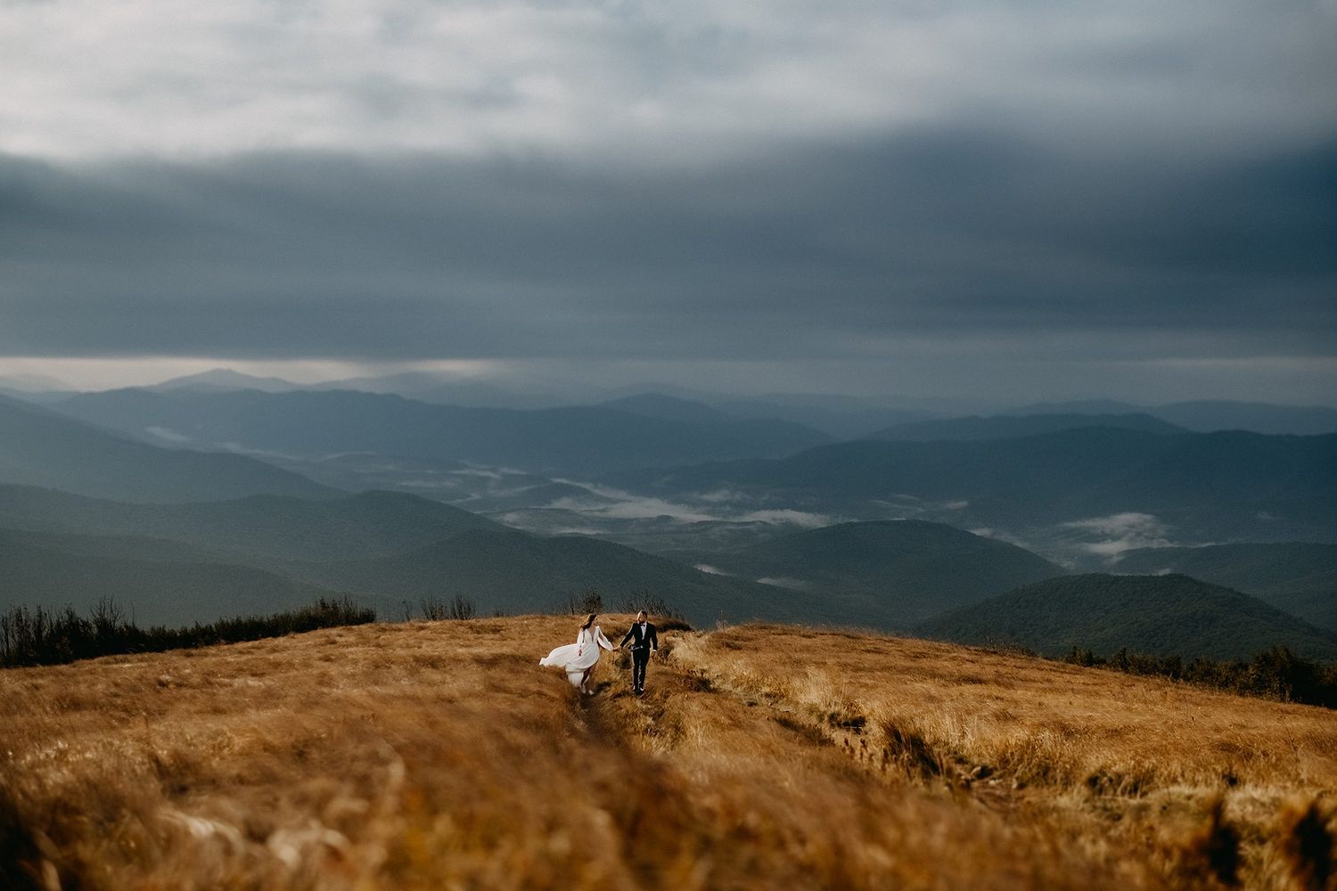 Bride and groom walking on a golden mountain meadow under dramatic storm clouds with misty valley views.