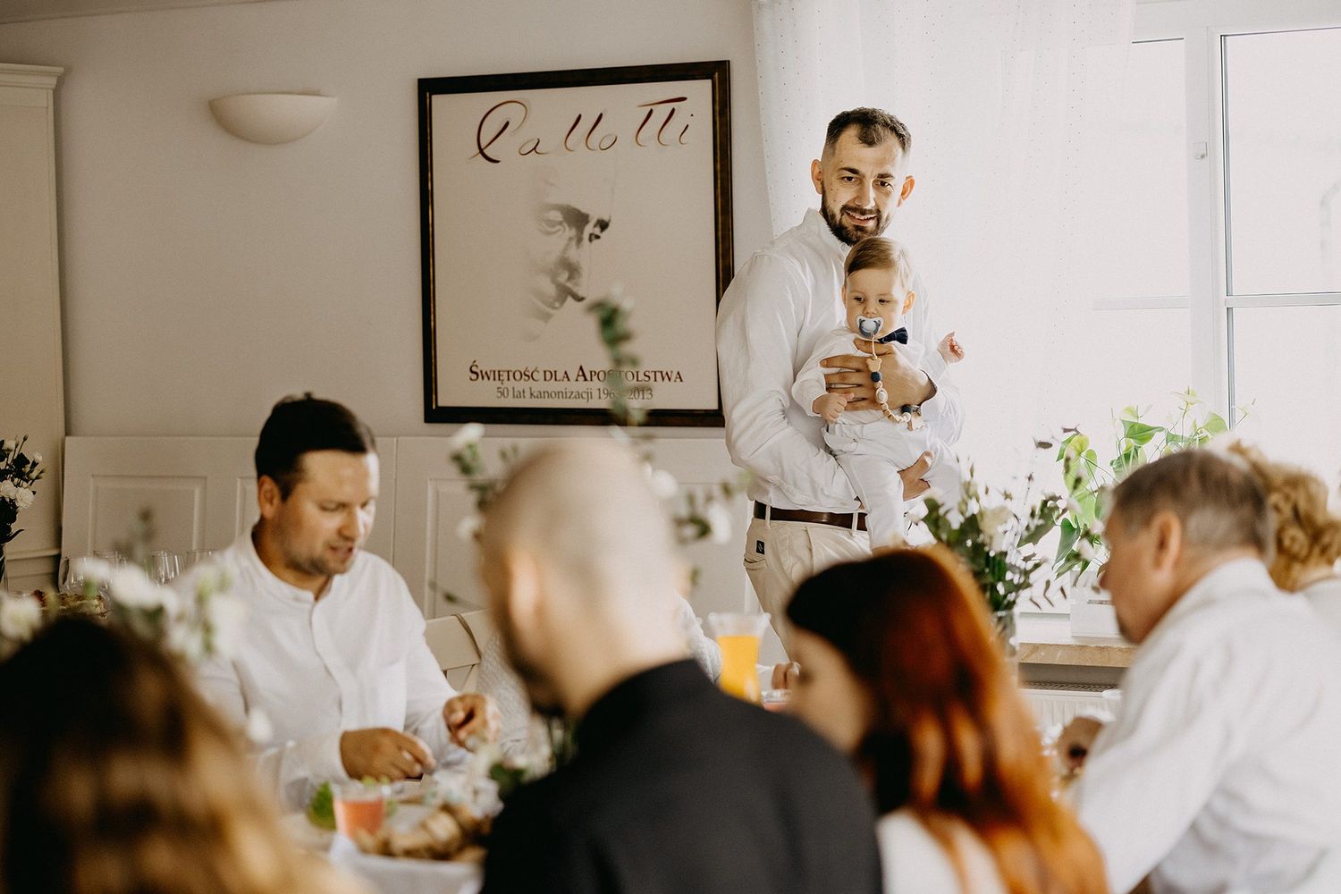 Man holding baby gives speech at elegant white-decorated dinner reception while guests dine.