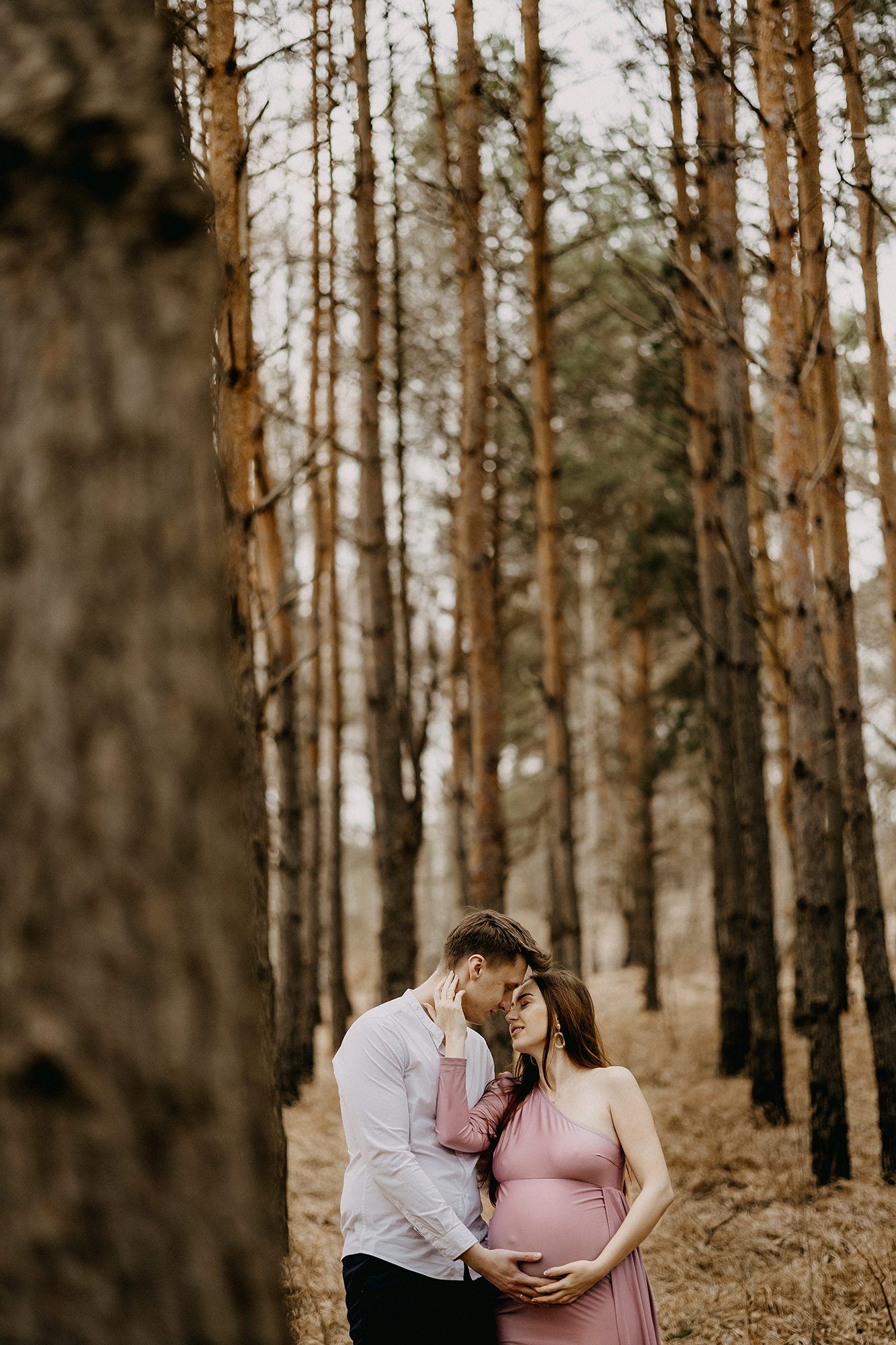 Expectant couple embracing tenderly in a pine forest, man kissing womans head in pink maternity dress.