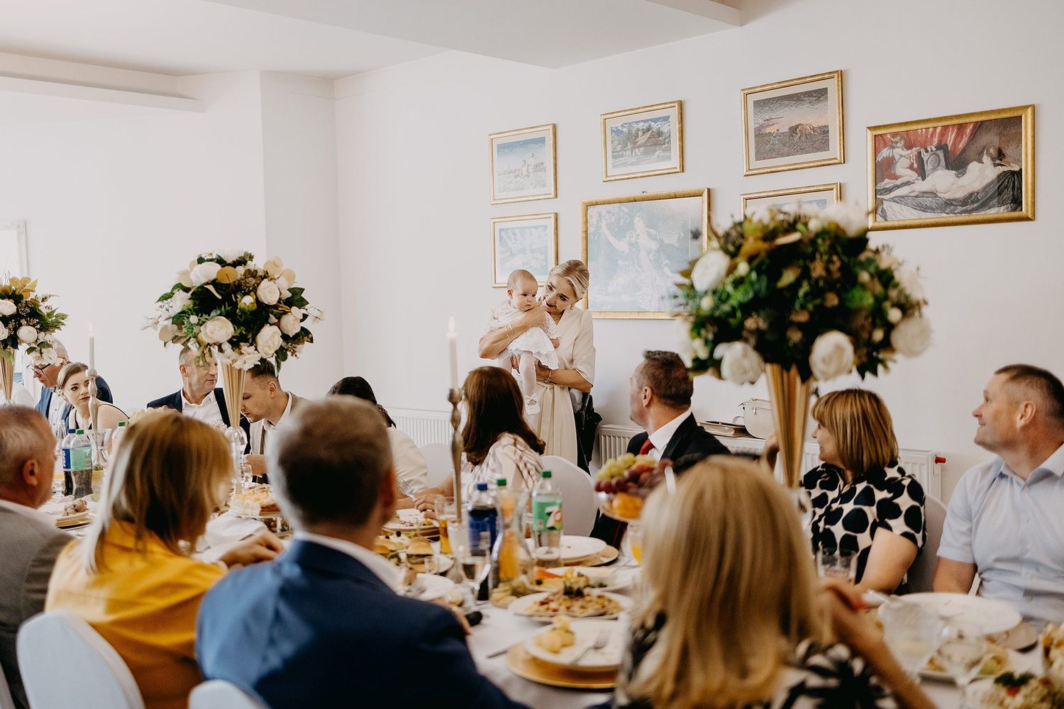 Guests gathered around decorated tables at an elegant indoor christening reception with floral centerpieces.