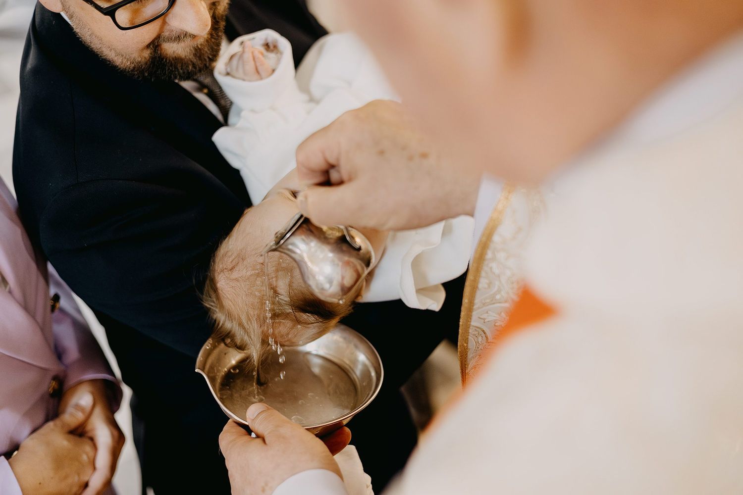 A priest pours holy water during a baptism ceremony with hands holding a silver bowl.