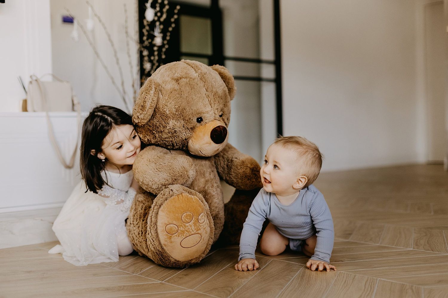 Toddler girl in white dress and baby boy sitting on floor with large teddy bear in bright room.