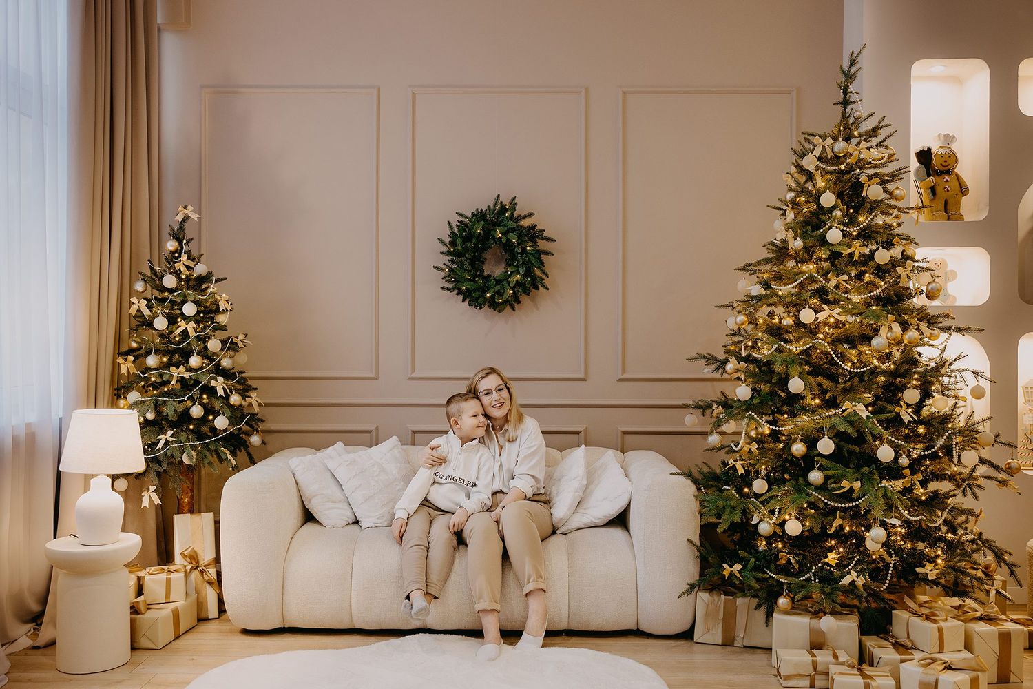 A mother and child sit together on a white sofa surrounded by elegantly decorated Christmas trees and wrapped gifts.