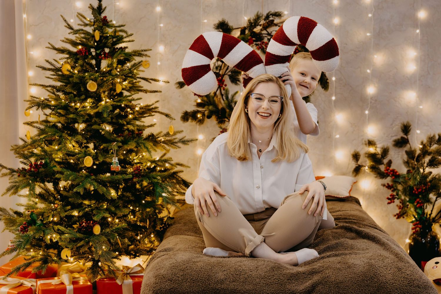 Smiling woman sits cross-legged on a cozy bed beside a lit Christmas tree while a child holds candy cane pinwheels above her head.