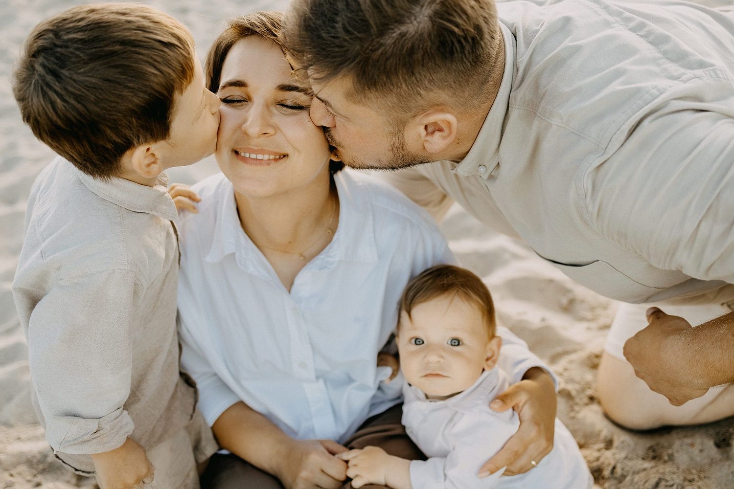 Happy mother receiving kisses from husband and son while baby sits on sandy beach at sunset.