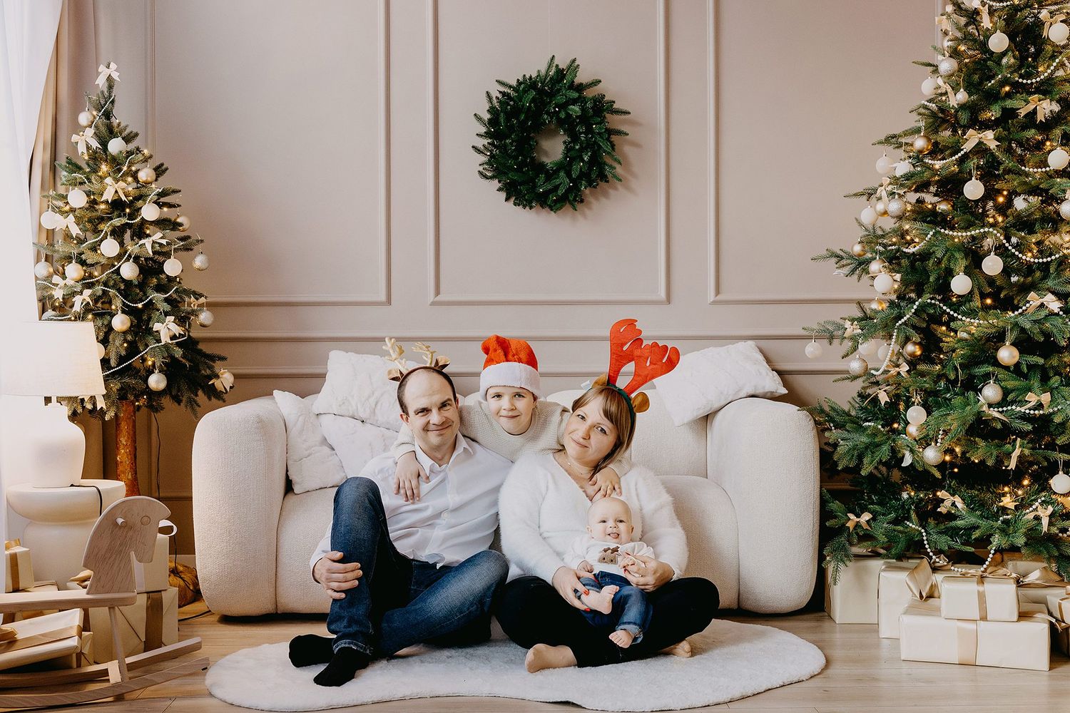 A smiling family of four wearing reindeer antlers sits together on the floor between two decorated Christmas trees.