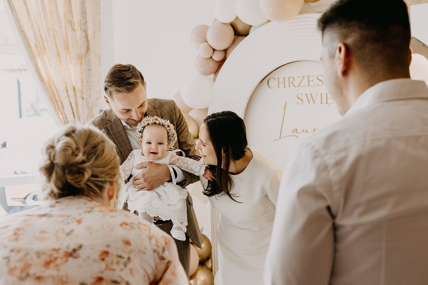 Parents with smiling baby at christening celebration near decorative baptism sign.